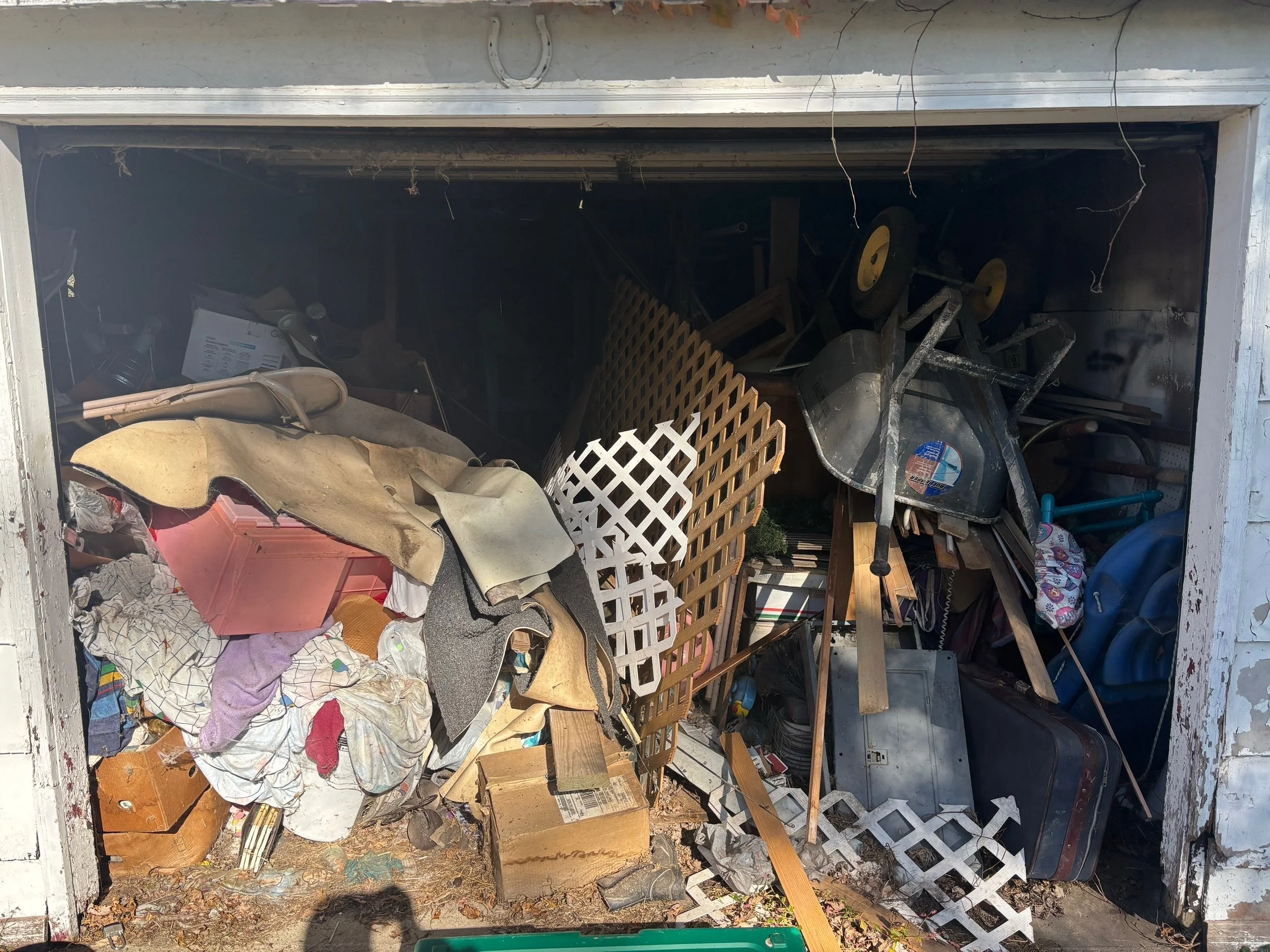 A cluttered garage filled with various items including a wheelbarrow, plastic lattice panels, a rolling suitcase, a blue children's bike, and piles of debris and miscellaneous objects.