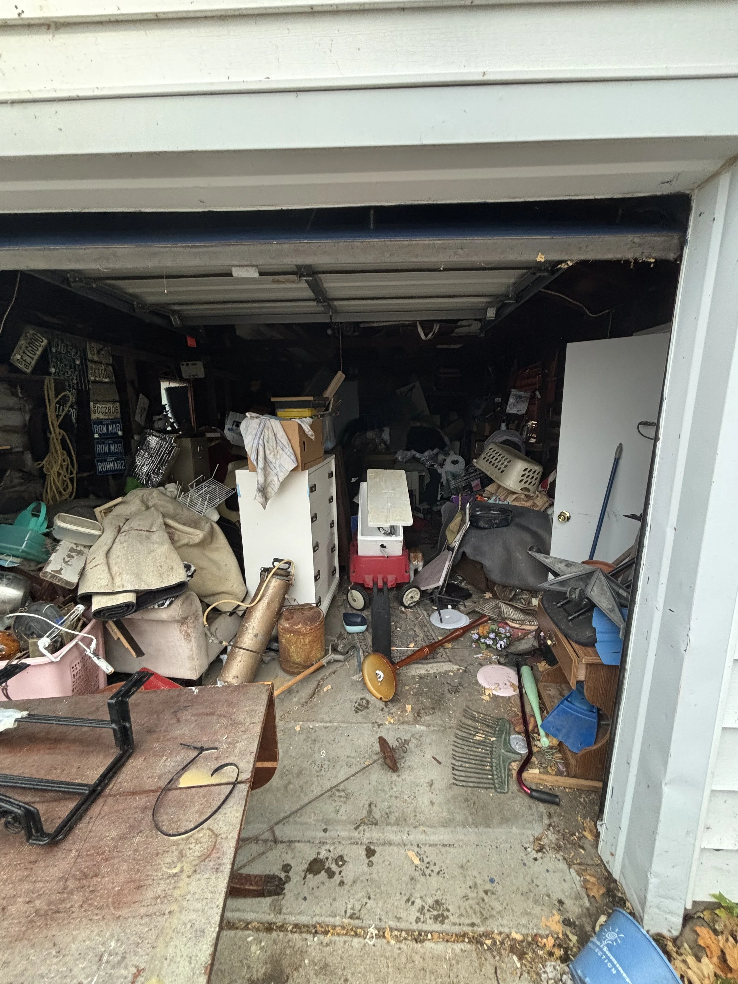 A cluttered garage filled with various household and gardening items, including a basket, a vacuum cleaner, tools, and boxes. The garage door is open.