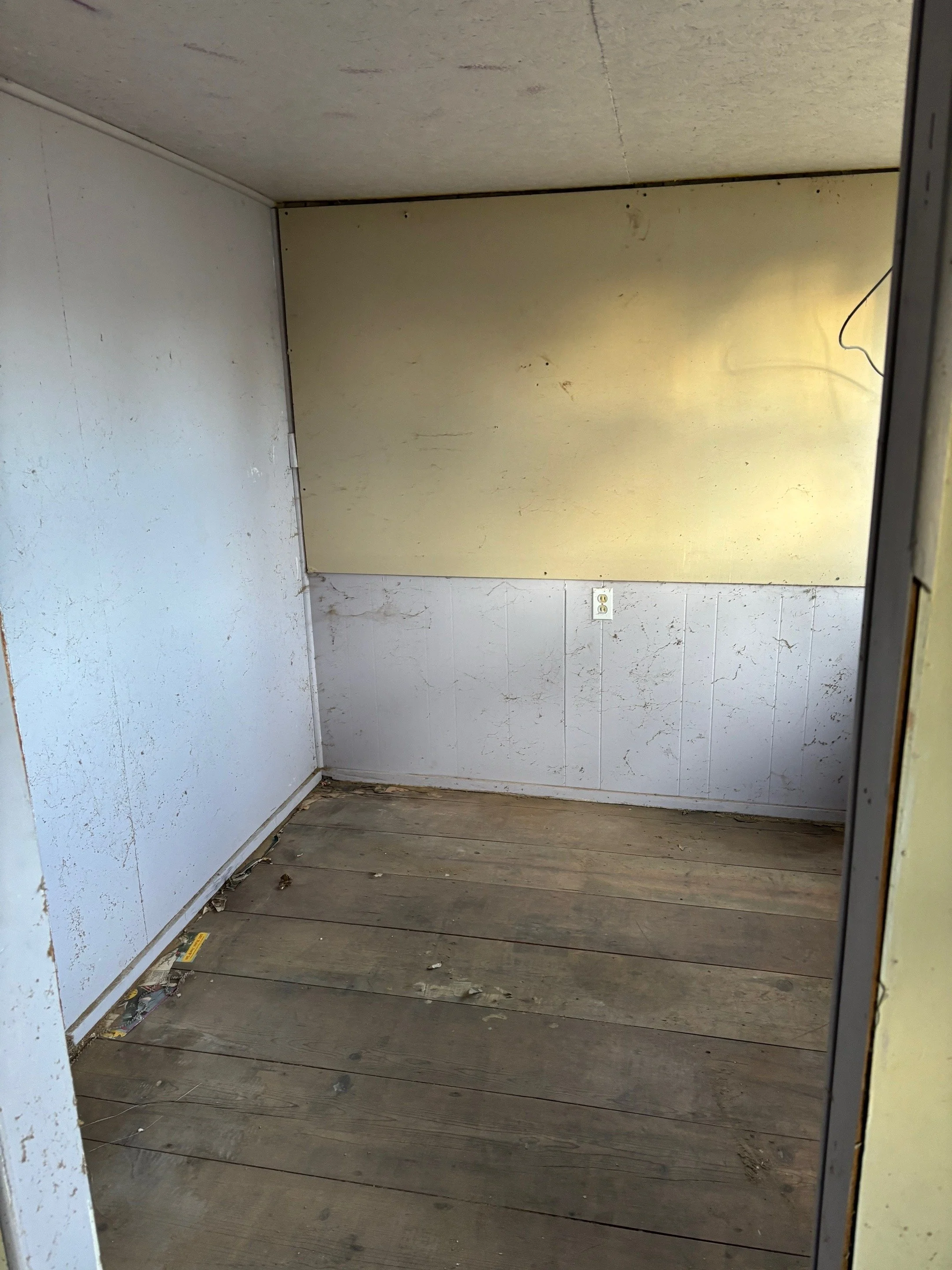 Empty small room with white walls, a yellow panel on the upper wall, an electrical outlet, and a wooden floor with dirt and debris.