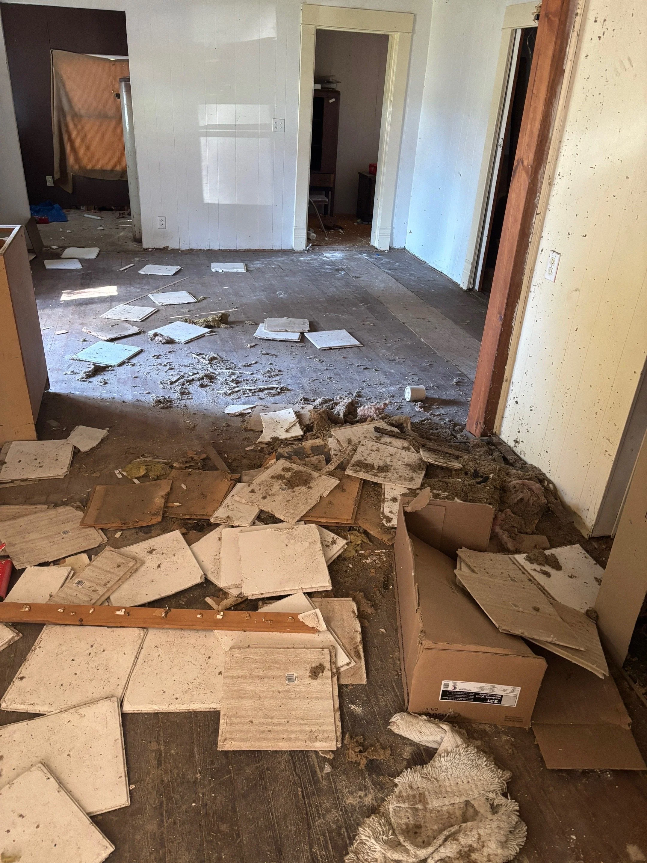 Disorganized and dirty room with broken tiles, debris, and cardboard boxes on the floor.