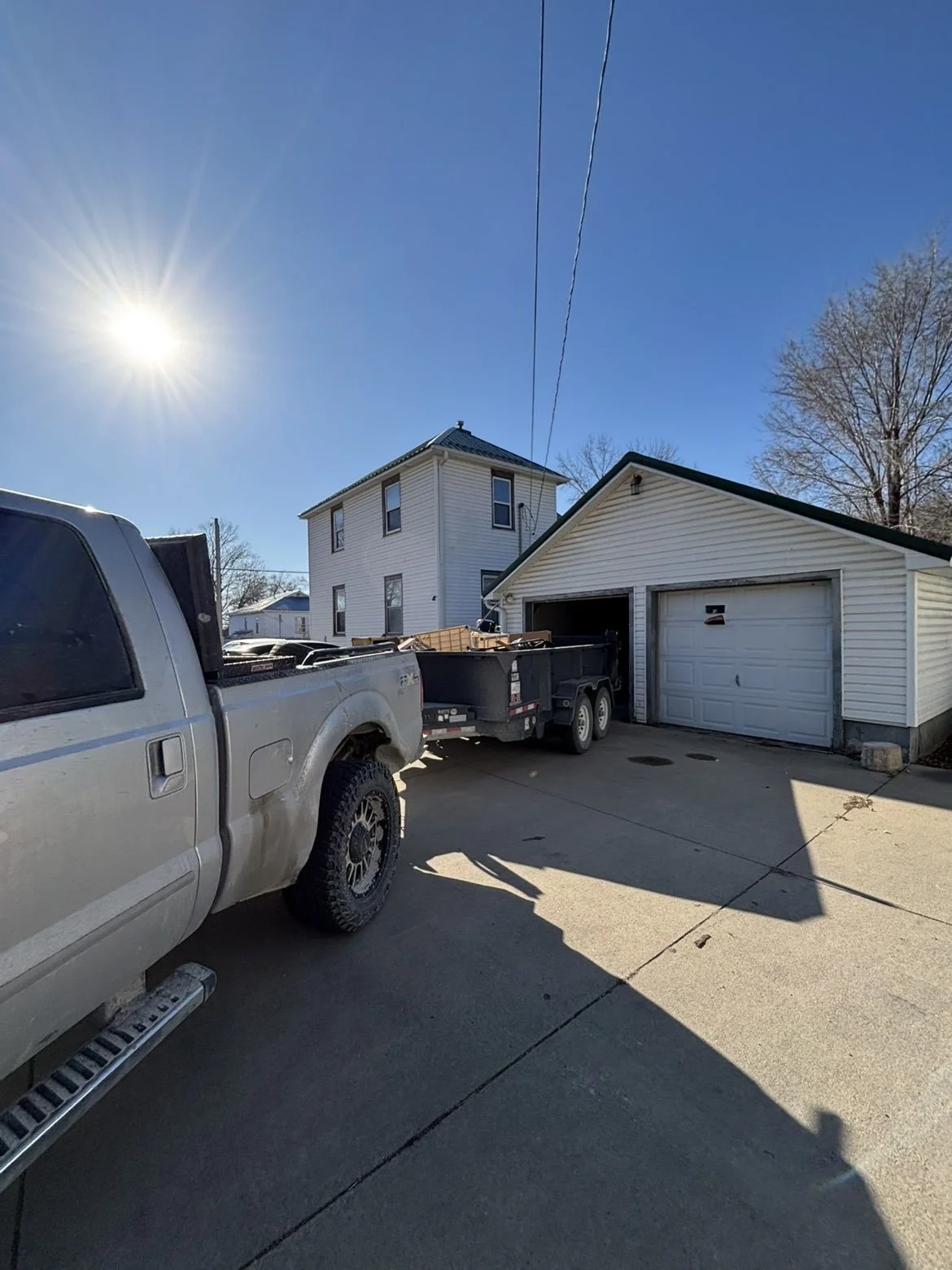 A residential driveway with a white truck towing a trailer filled with wood and other materials, next to a garage. The house in the background is a two-story structure under a clear blue sky with the sun shining.