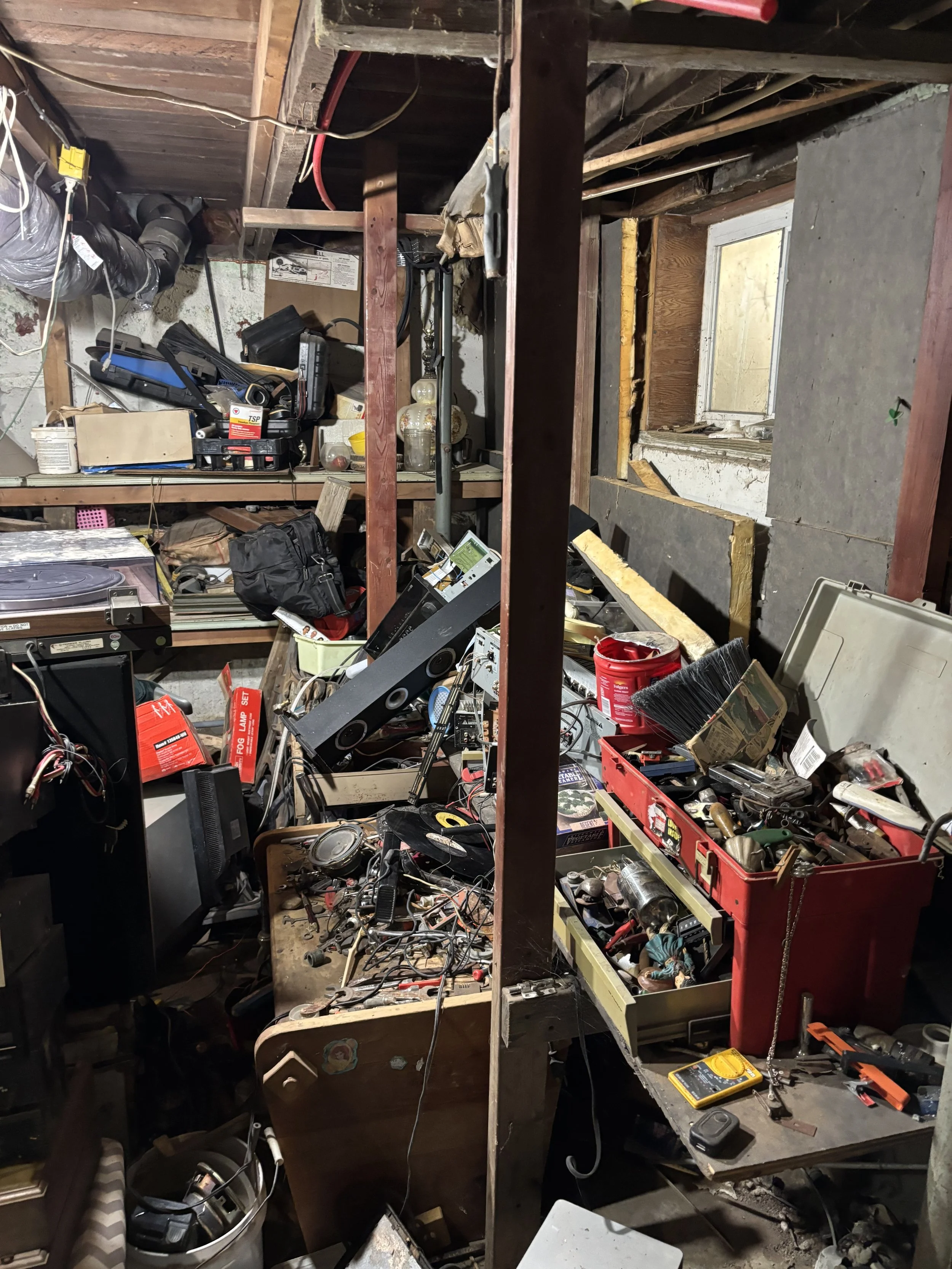 A cluttered workshop or garage with shelves filled with tools, equipment, and miscellaneous items. The workspace appears disorganized with various tools, wires, and objects scattered around.