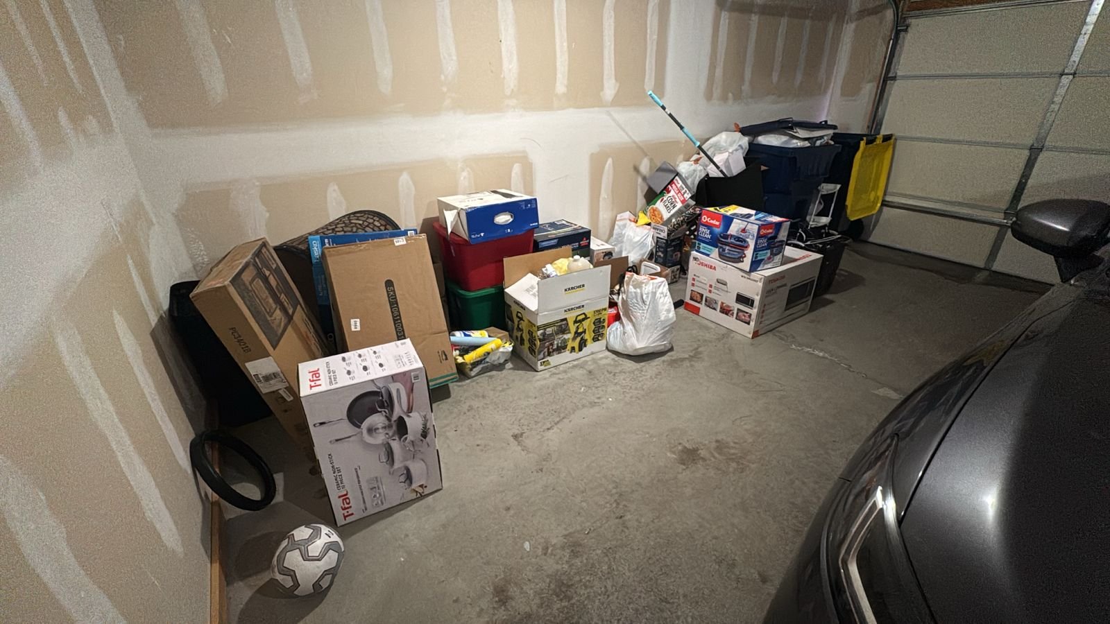 Garage with various boxes and items stacked along the wall and on the floor, including a soccer ball, a boxed air fryer, and some yard tools. Part of a black vehicle is visible at the right edge.