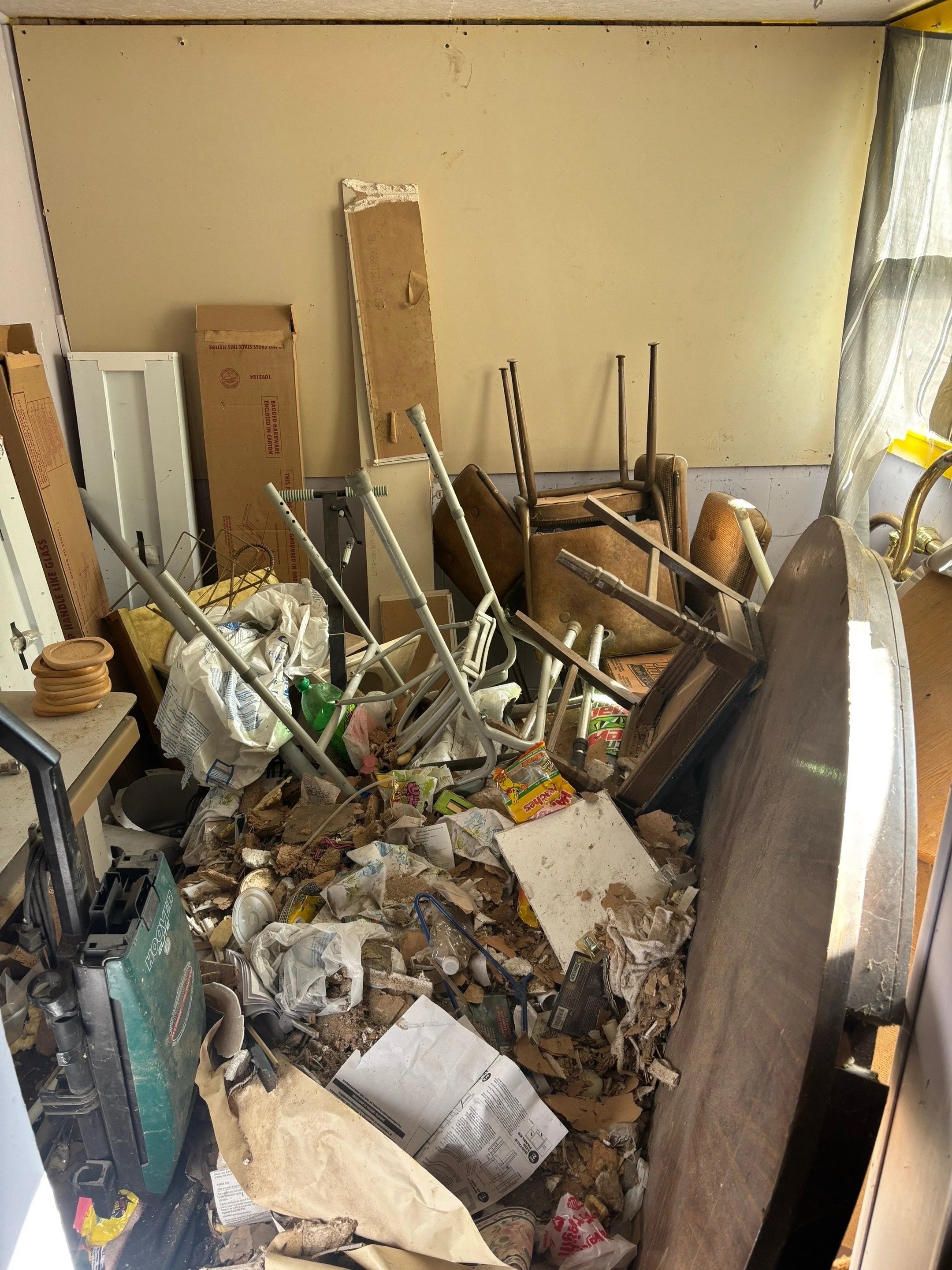 Cluttered room with toppled chairs, scattered trash, boxes, and debris on the floor, and a cluttered table on the left.