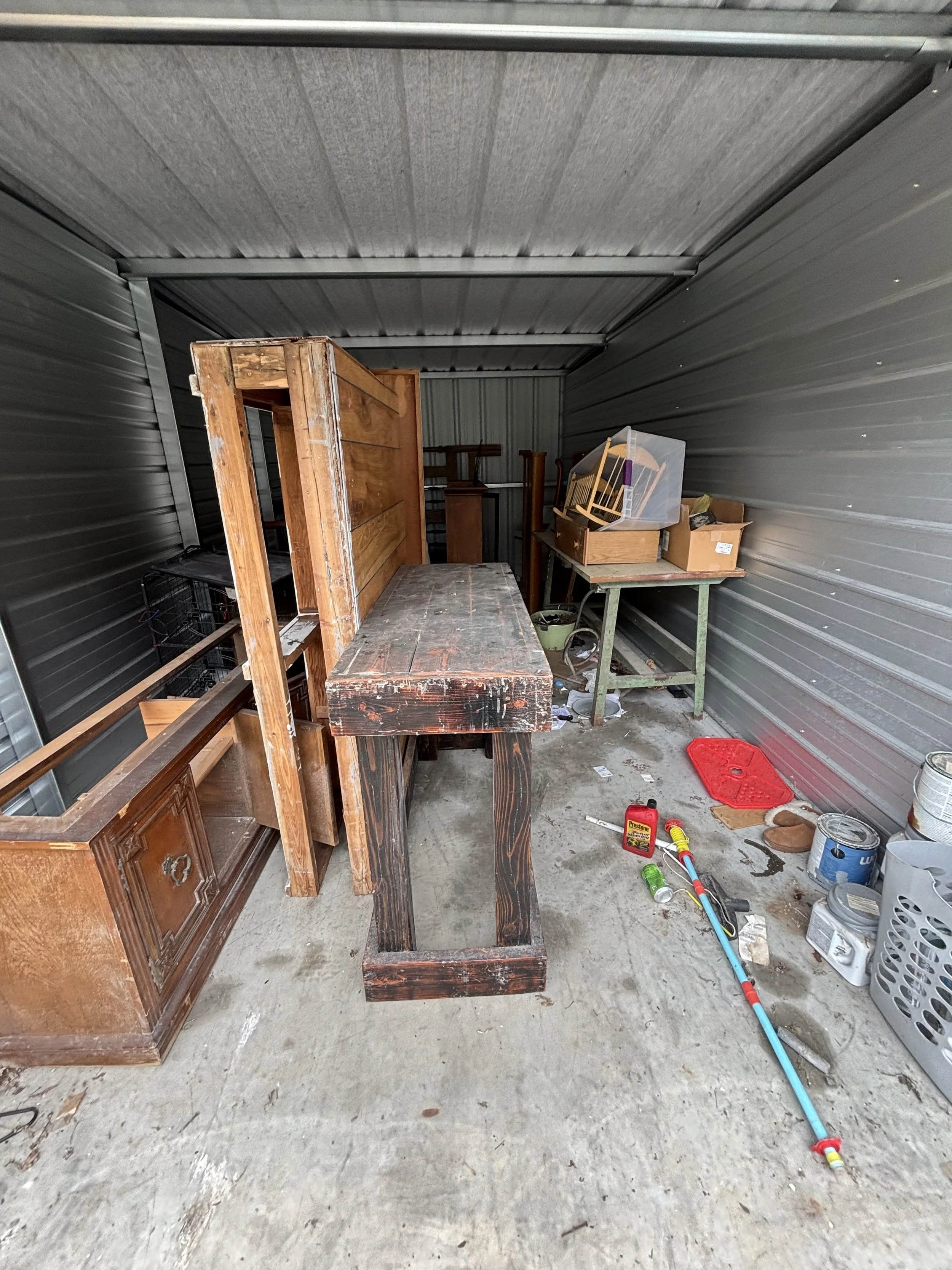 Storage unit with various furniture, tools, and supplies including a wooden piece, a small table, a plastic container, cans, and a garden hose on the concrete floor.