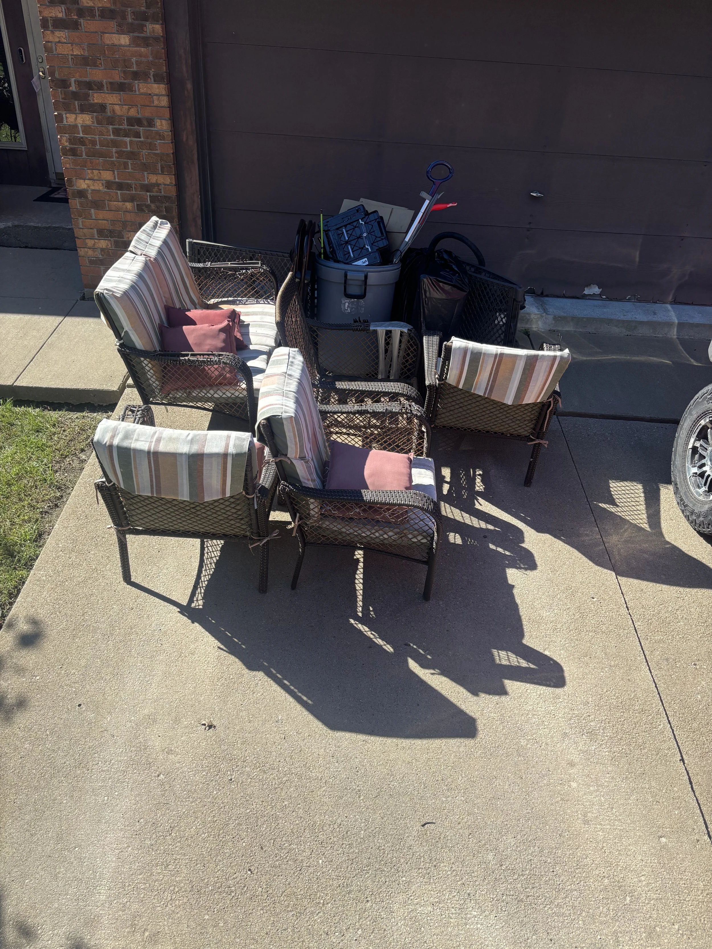 Outdoor patio scene with chairs, cushions, and a collection of tools and equipment against a dark brown garage door.