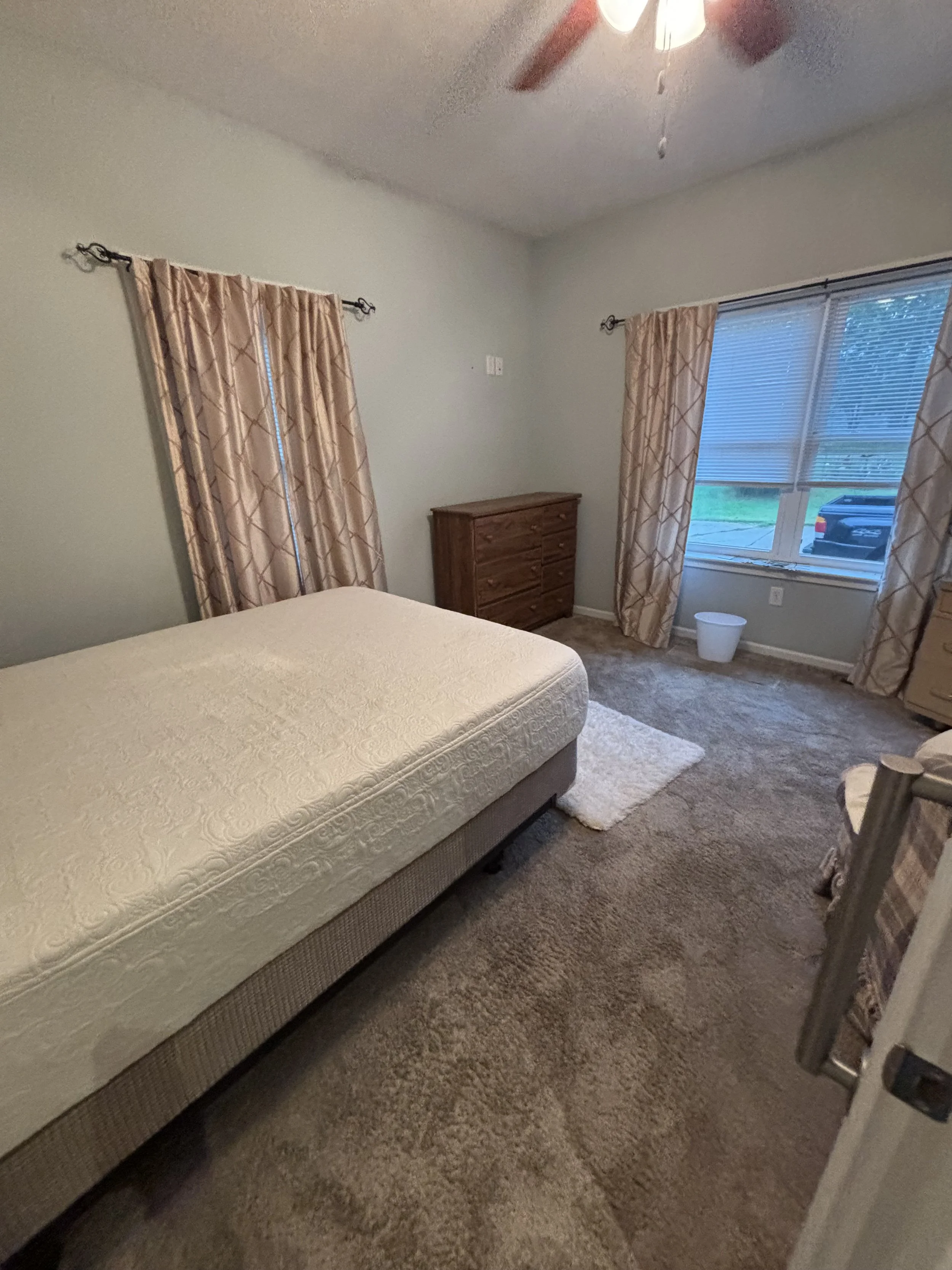 A bedroom with a bed that has a white mattress, a wooden dresser, beige curtains, a window with blinds, a beige carpet, and a ceiling fan with lights.
