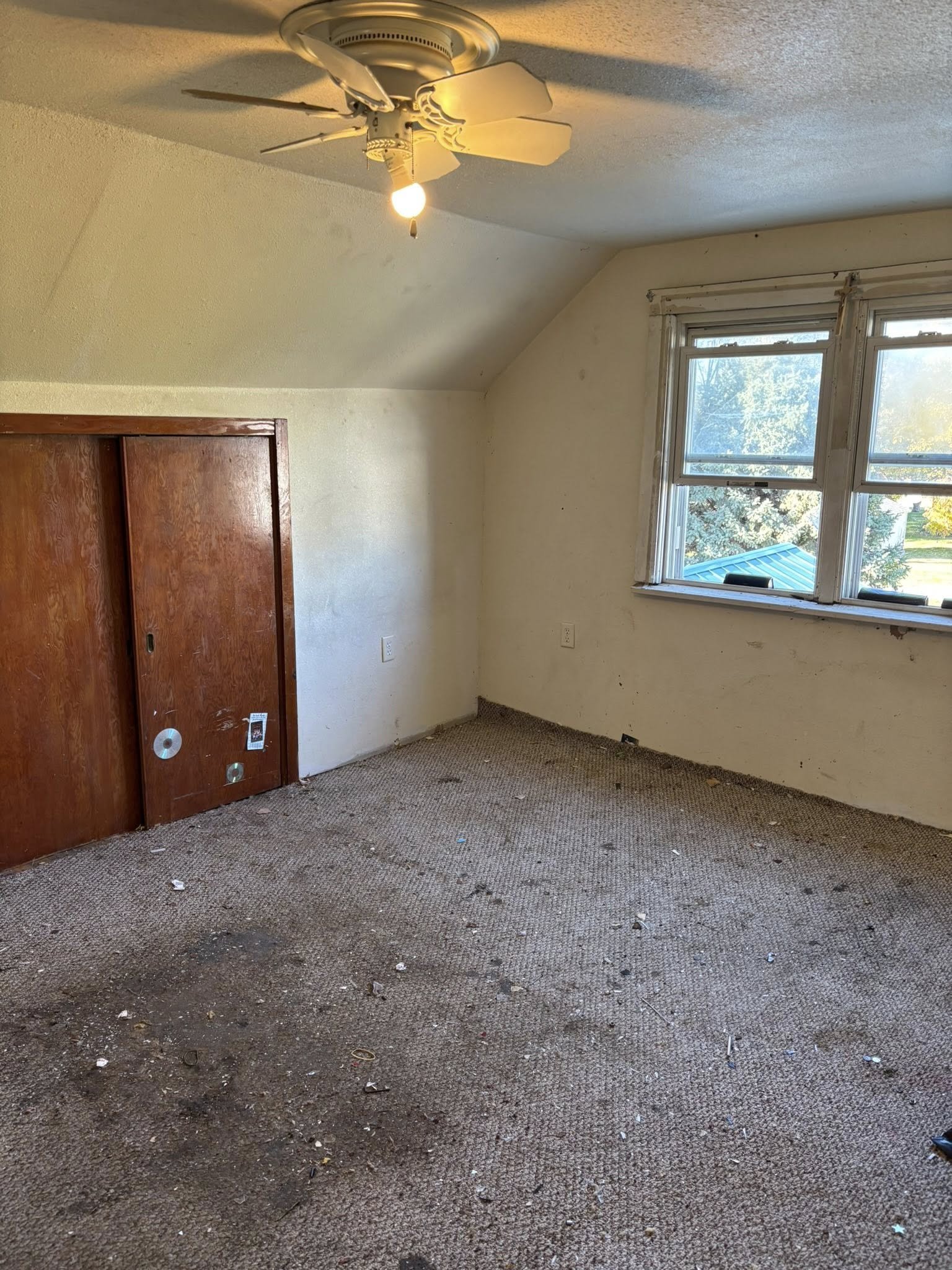 Empty, dirty room with stained carpet, a ceiling fan with a light bulb, and two windows showing an outdoor scene with trees and sky.