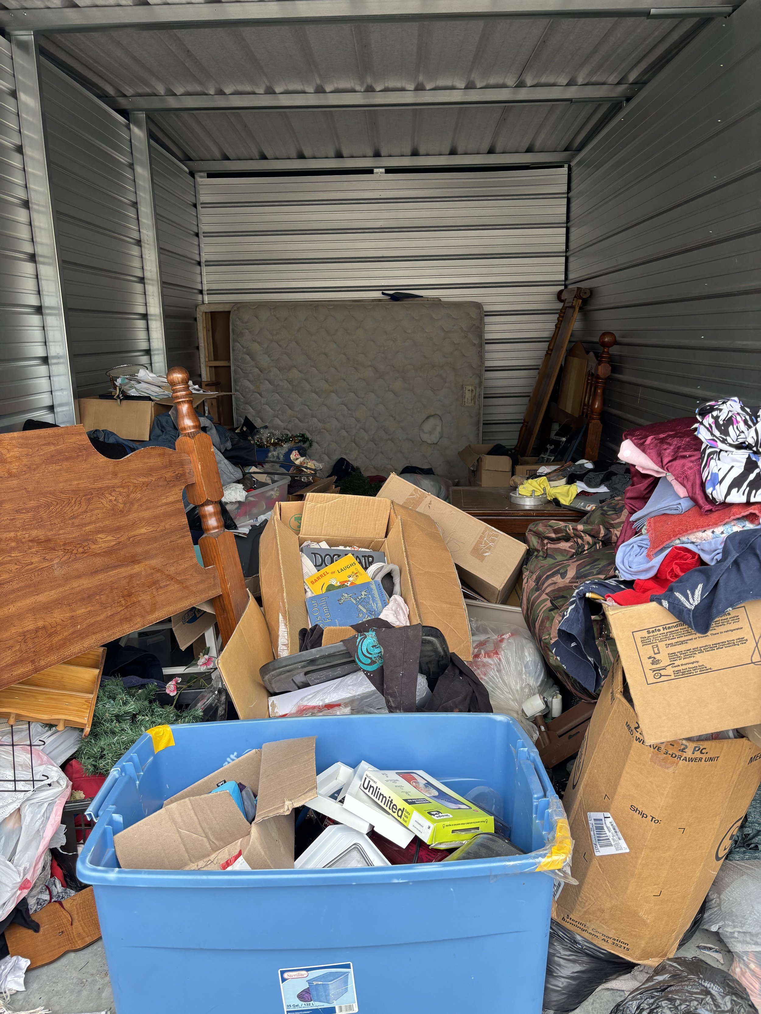 A cluttered storage unit filled with various boxes, scattered clothes, a mattress, a wooden headboard, and a blue plastic storage bin.