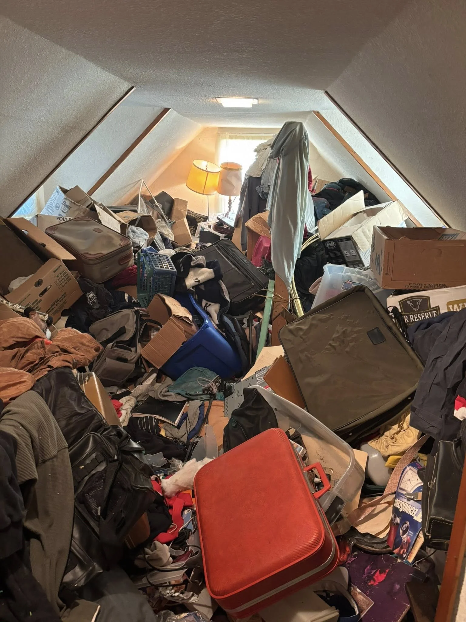 Cluttered attic with clothes, boxes, and suitcases, illuminated by natural light from a window at the far end.