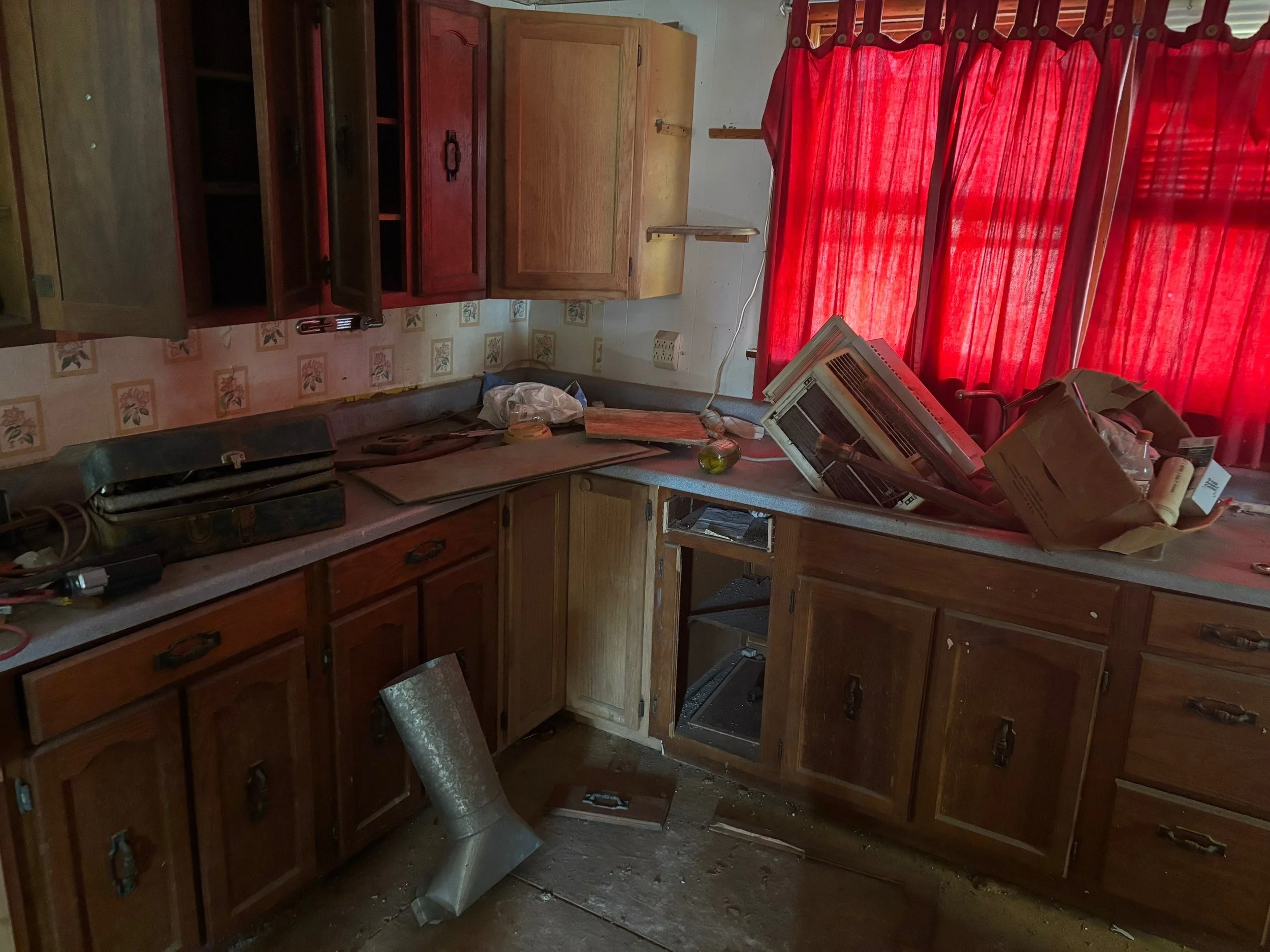 A cluttered kitchen with open cabinets, red curtains, and dusty surfaces, containing various items including a disorganized paper bag, a broken air conditioning unit, and a metal vent on the floor.