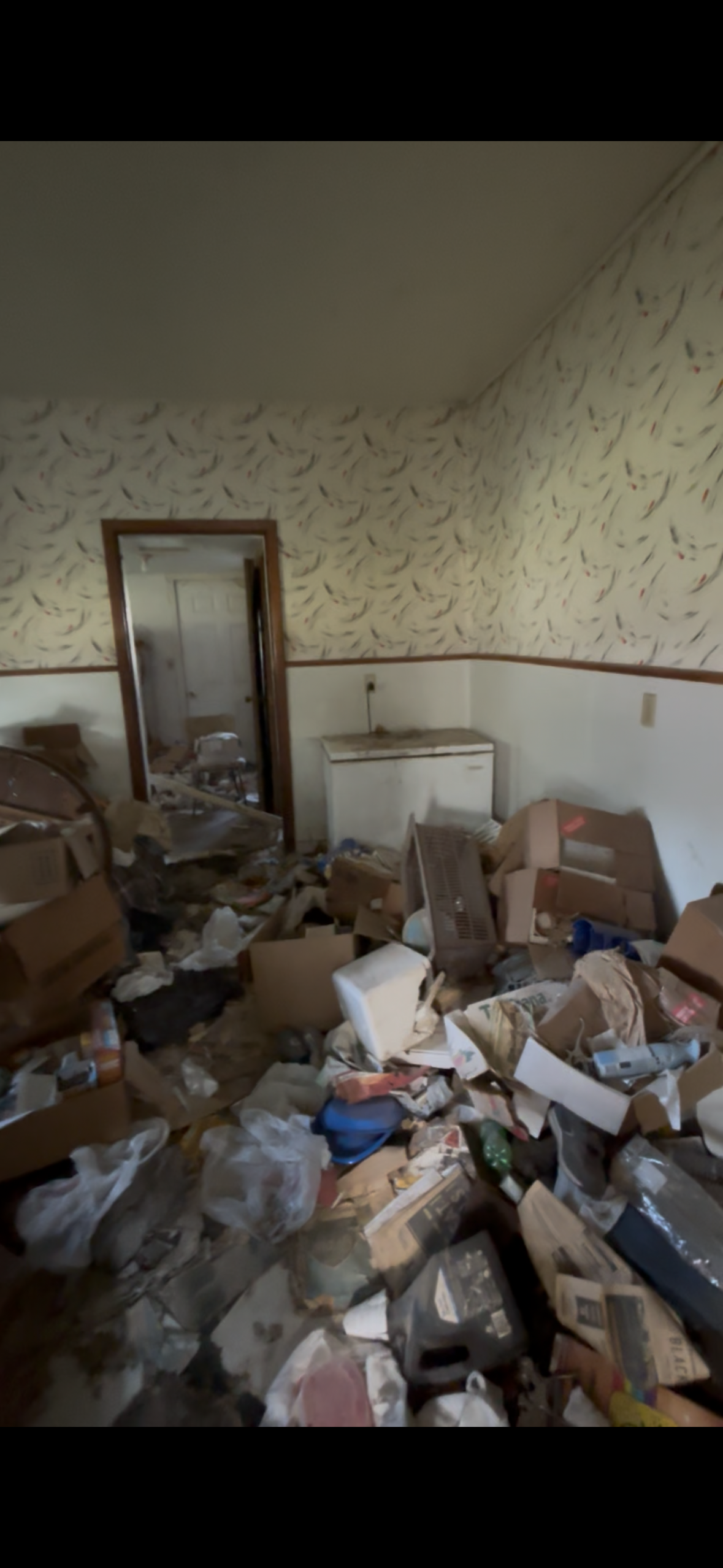 A cluttered room filled with trash, cardboard boxes, and debris, with wallpapered walls and an open doorway leading to another messy space.