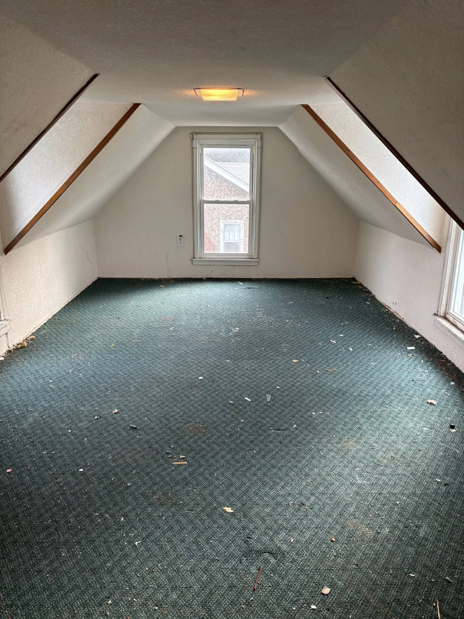 Empty, unfinished attic room with sloped ceilings, white walls, a small window, and green carpet with debris.