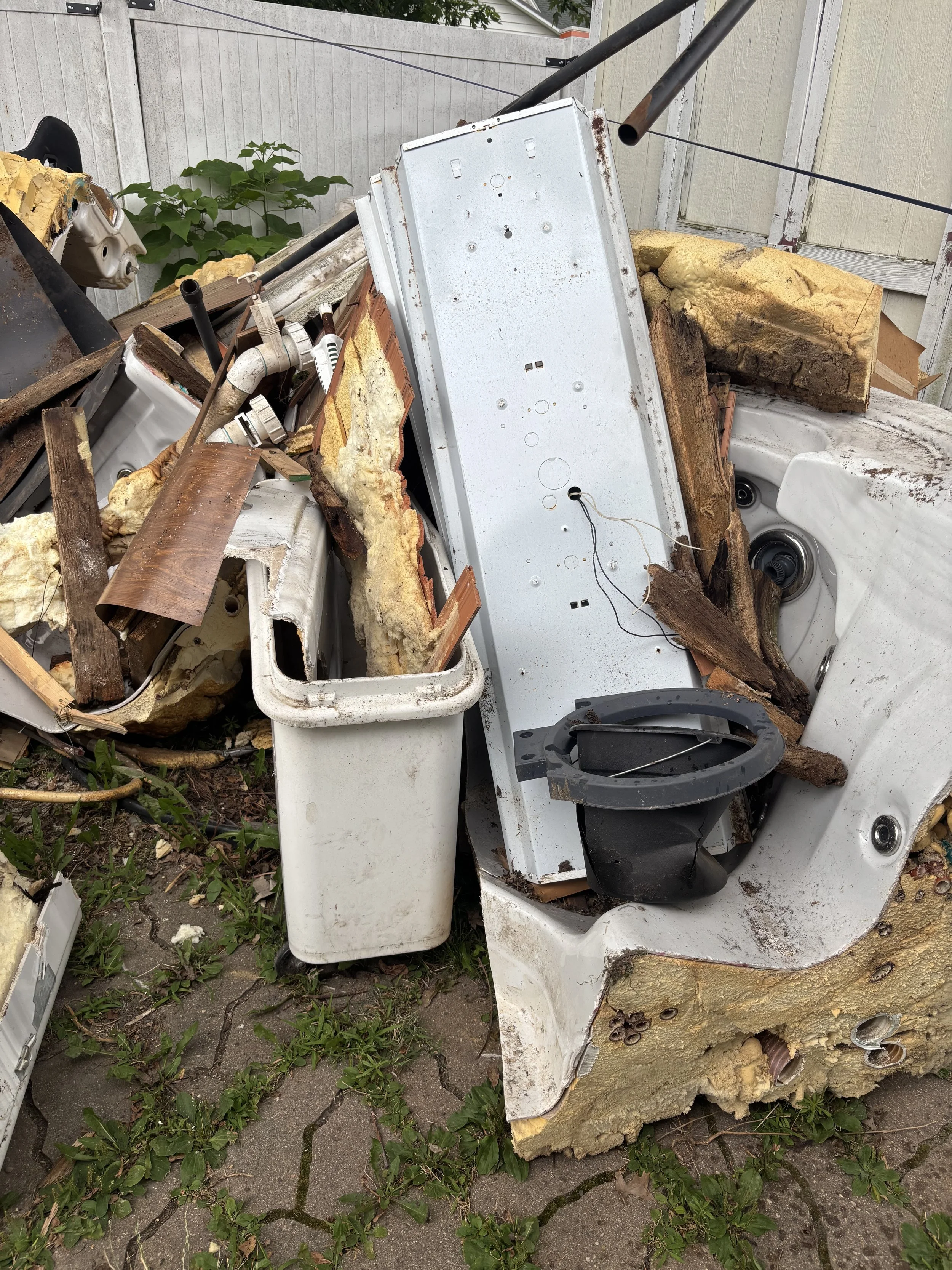 A pile of discarded household appliances including a washing machine and a dryer, with yellow insulation foam, wood, pipes, and some greenery growing among the debris outside near a weathered fence.