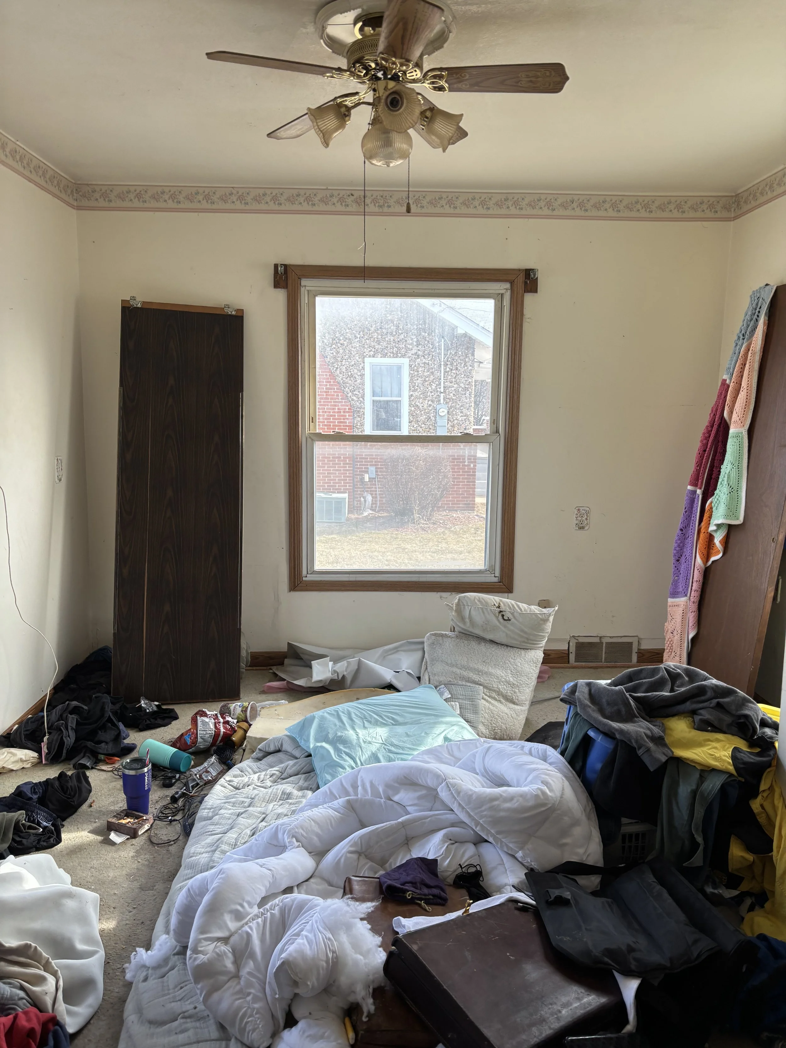 Disorganized bedroom with bedding on the floor, clothes and miscellaneous items scattered, a window showing a brick house outside, and a ceiling fan with lights.