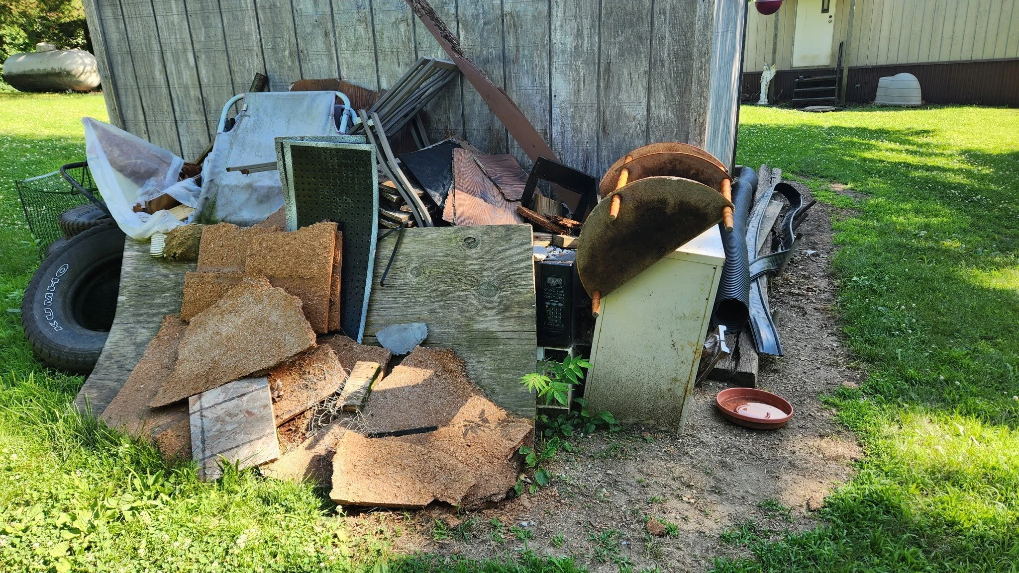 Piles of discarded objects and debris outside a shed, including bricks, metal, wood, a microwave, a small plant, a tire, a plastic bowl, and various miscellaneous items.