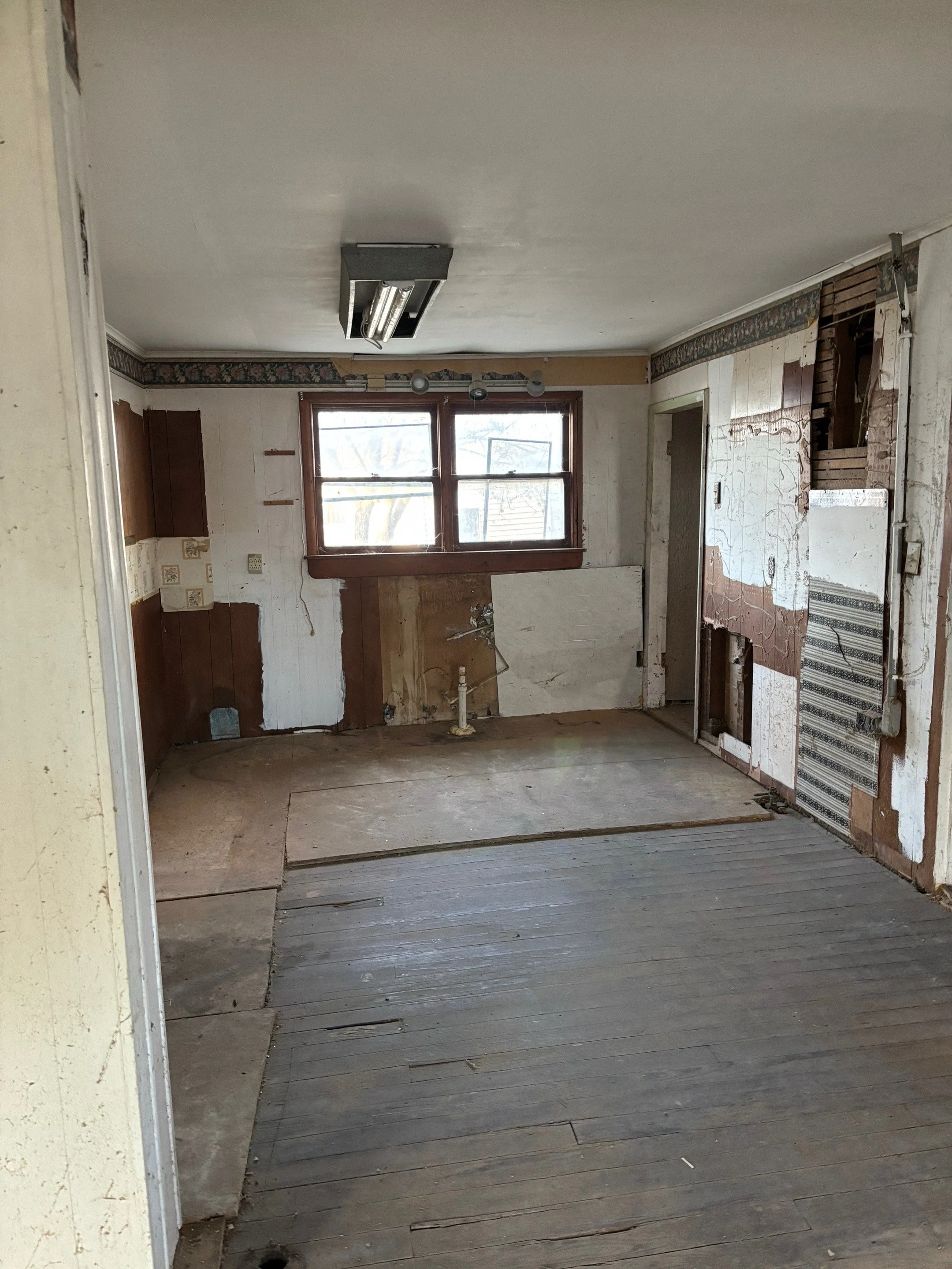 Room with damaged walls and flooring, raw wood and exposed areas, window at the back, and visible plumbing pipes.