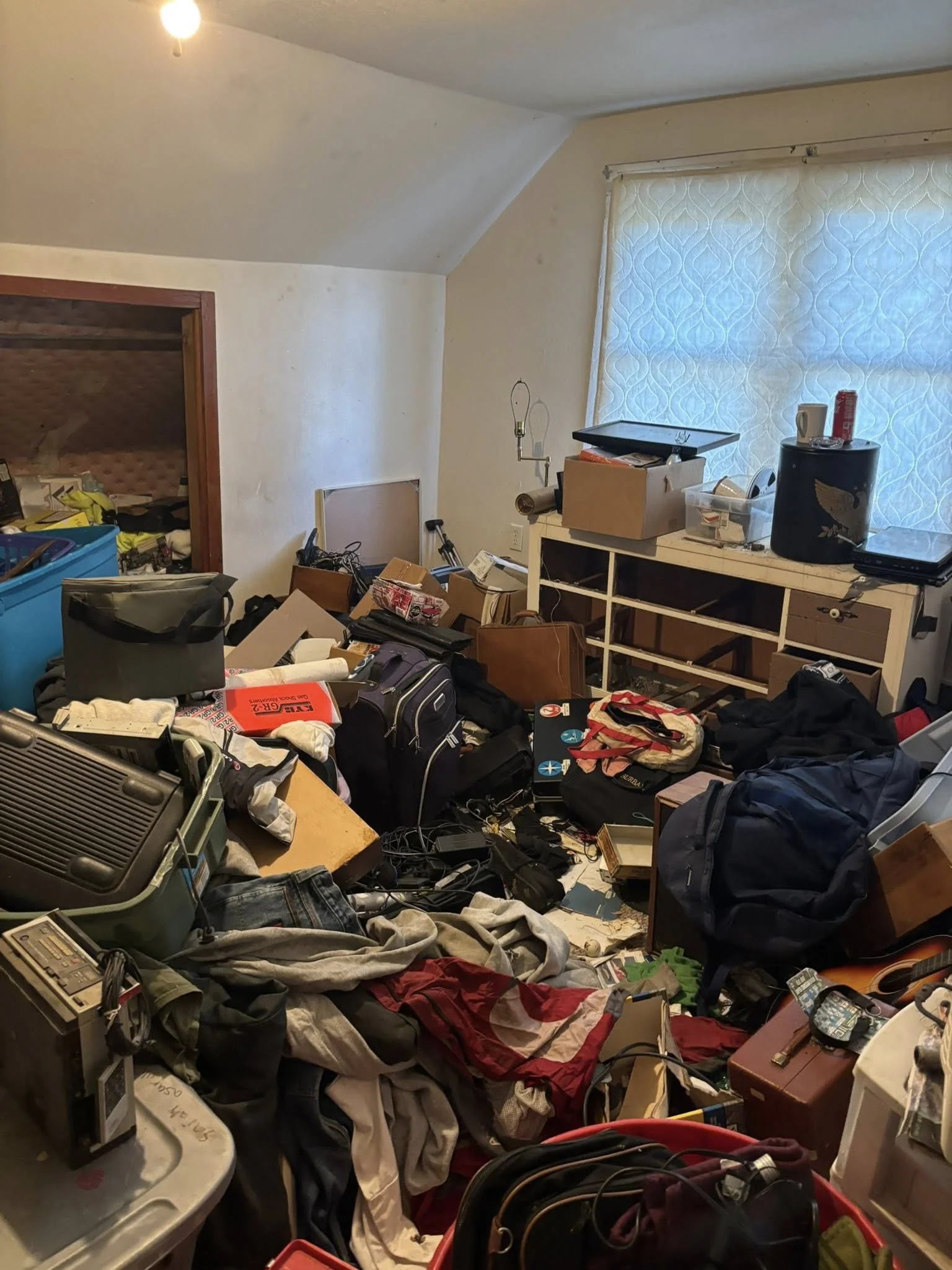 Cluttered room with various items including clothes, boxes, bags, and electronic devices scattered across the floor; a large window with a white curtain, and a small shelf or cabinet against the wall.