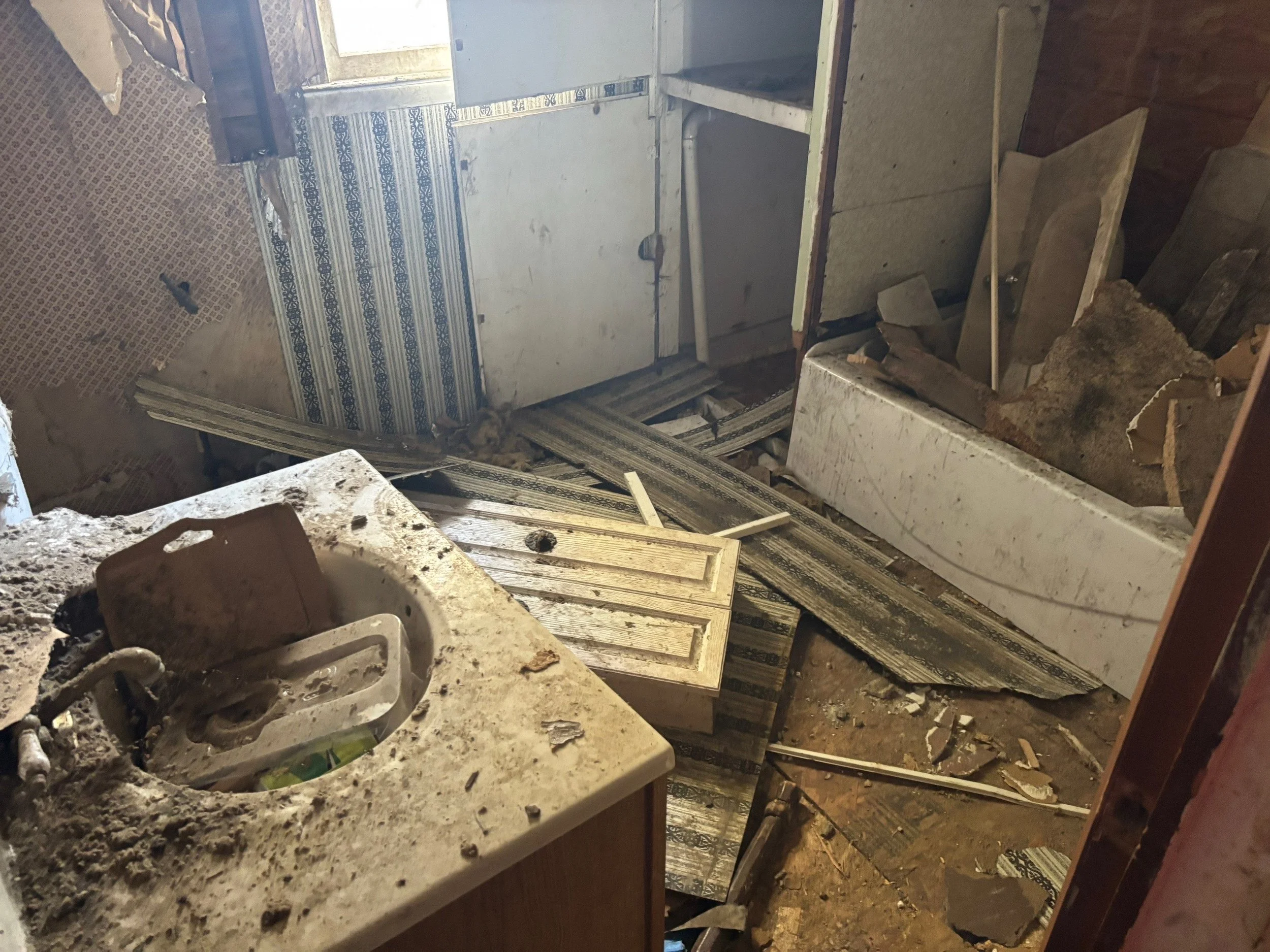 Dilapidated bathroom with a broken sink, damaged walls, a dirty bathtub, scattered debris, and damaged flooring.