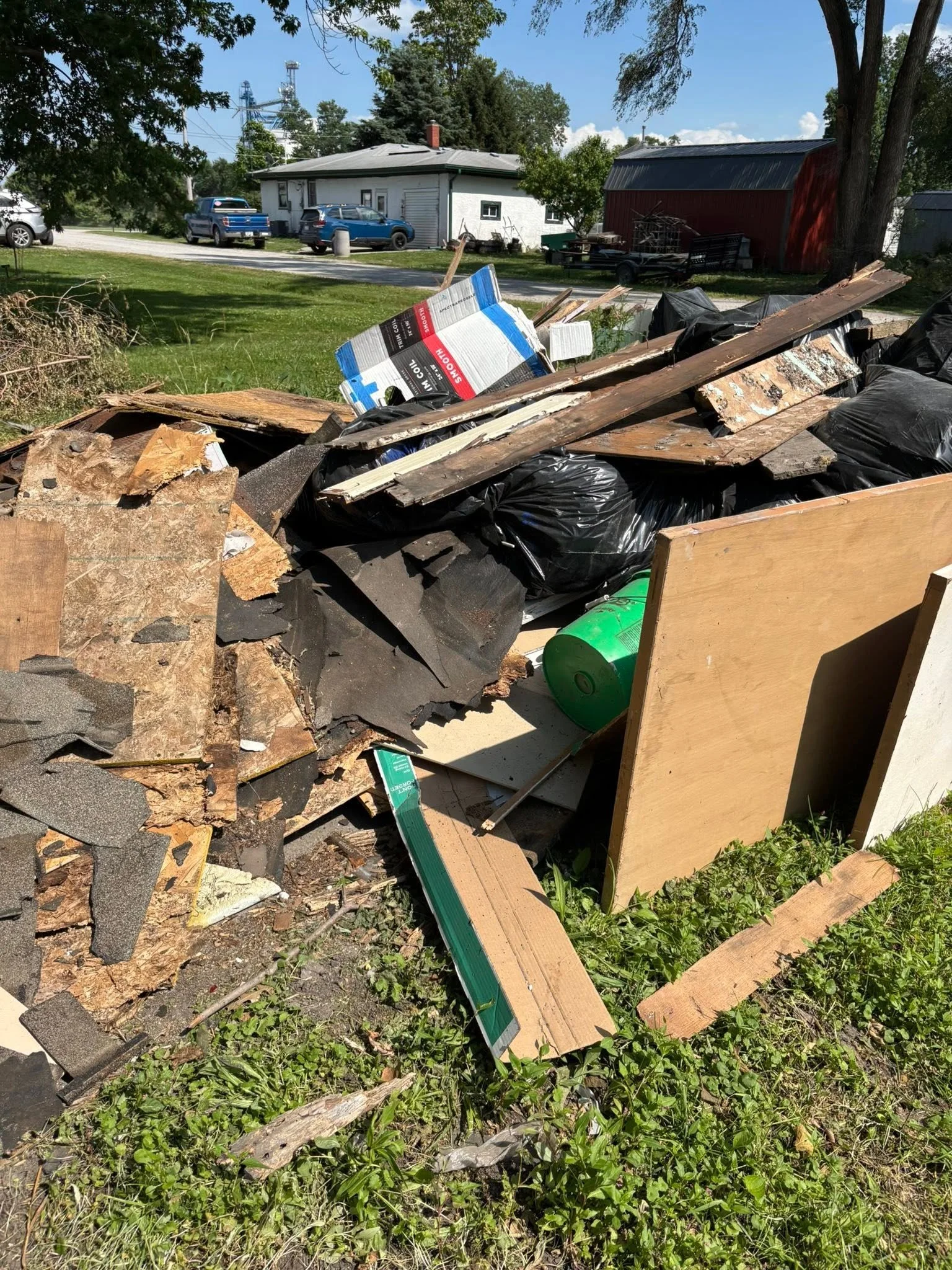 Piles of construction debris, including wood, black trash bags, and a green plastic container, outside on the grass with buildings and vehicles in the background.