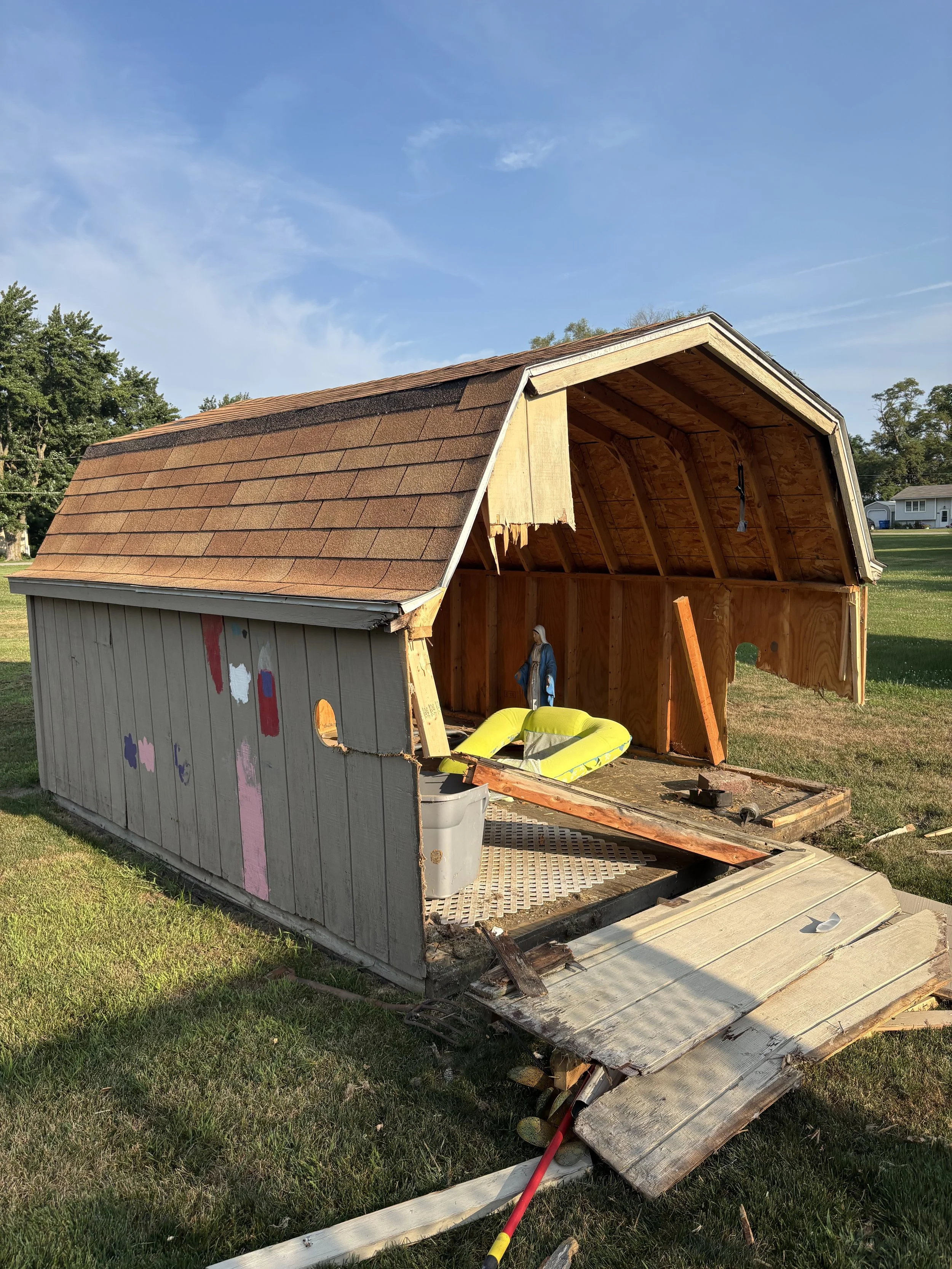 A small wooden shed with part of its roof and walls damaged, exposing the interior. Debris and construction tools are on the ground, indicating recent destruction or renovation.