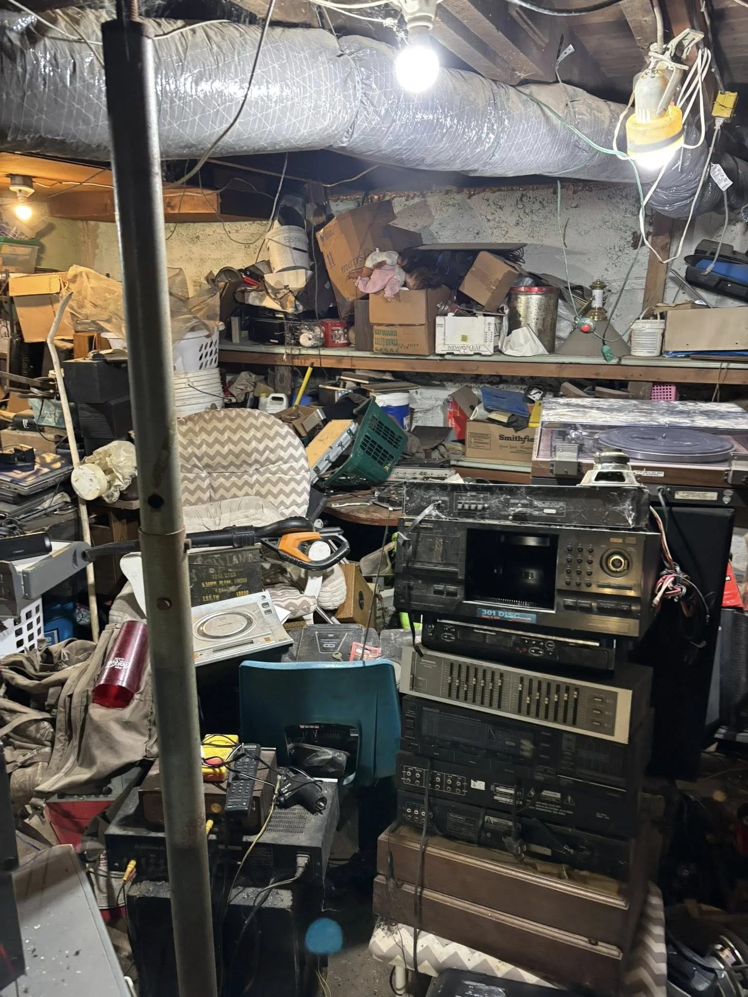 A cluttered basement with shelves filled with various objects, a stereo system, electronic equipment, boxes, and miscellaneous items. The ceiling has exposed pipes and insulation, with a hanging light bulb illuminating the space.