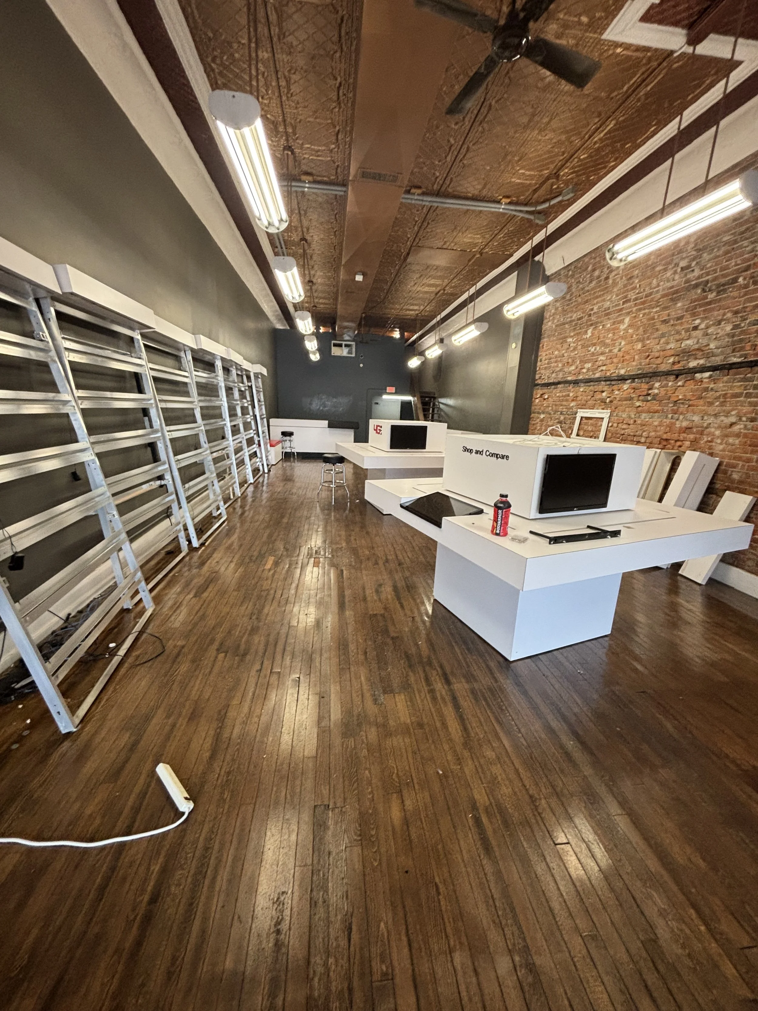 Empty retail or office space with dark wooden floors, exposed brick wall on the right, a dark gray wall at the end, and a ceiling with decorative metallic panels. There are white display counters with monitors, a black stool, a ladder, hanging fluorescent lights, and various items like a water bottle and power strip on the counters.