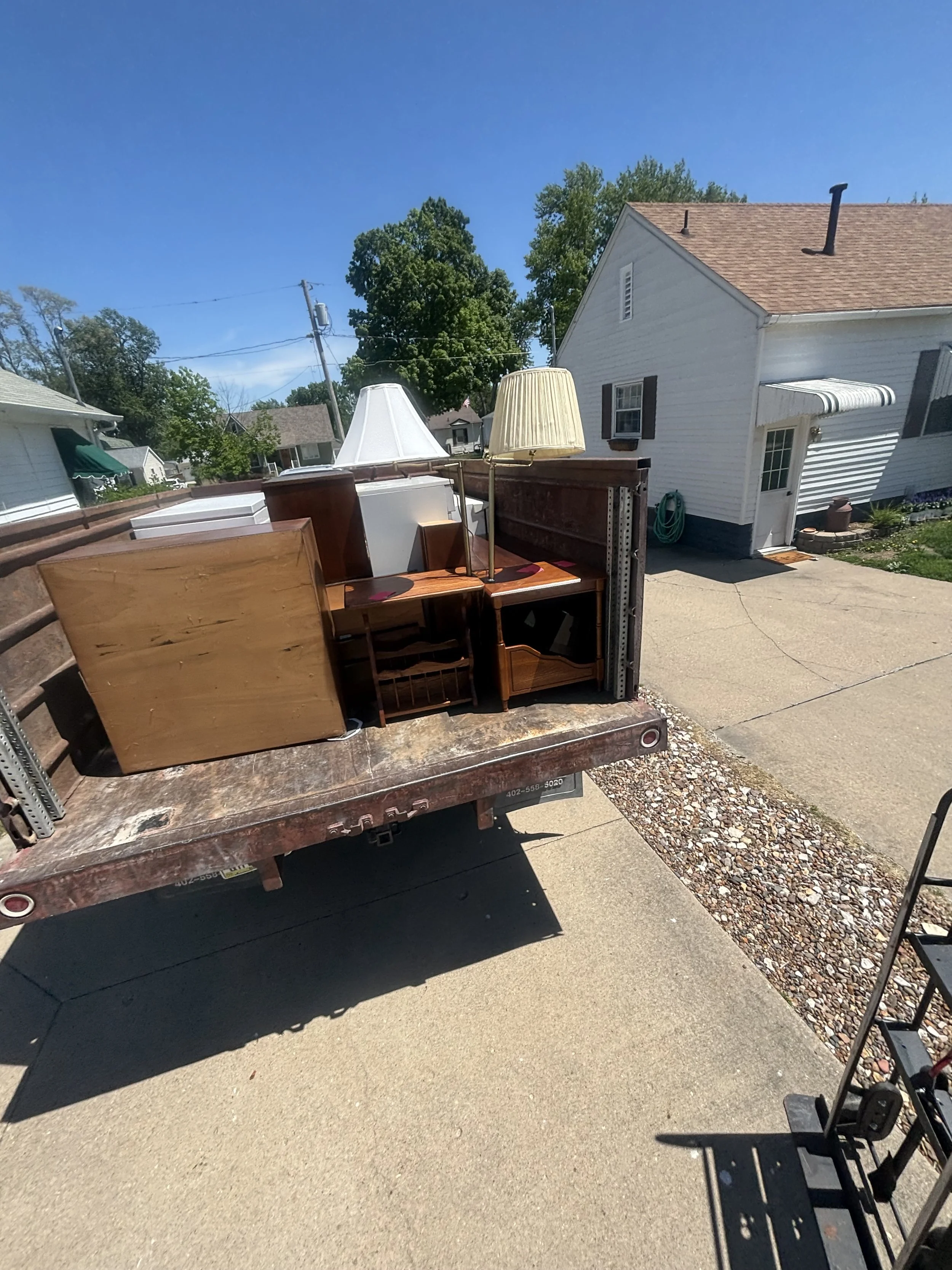 A truck carrying furniture, including lamps and a wooden cabinet, parked in a driveway next to a house with white siding and black shutters.