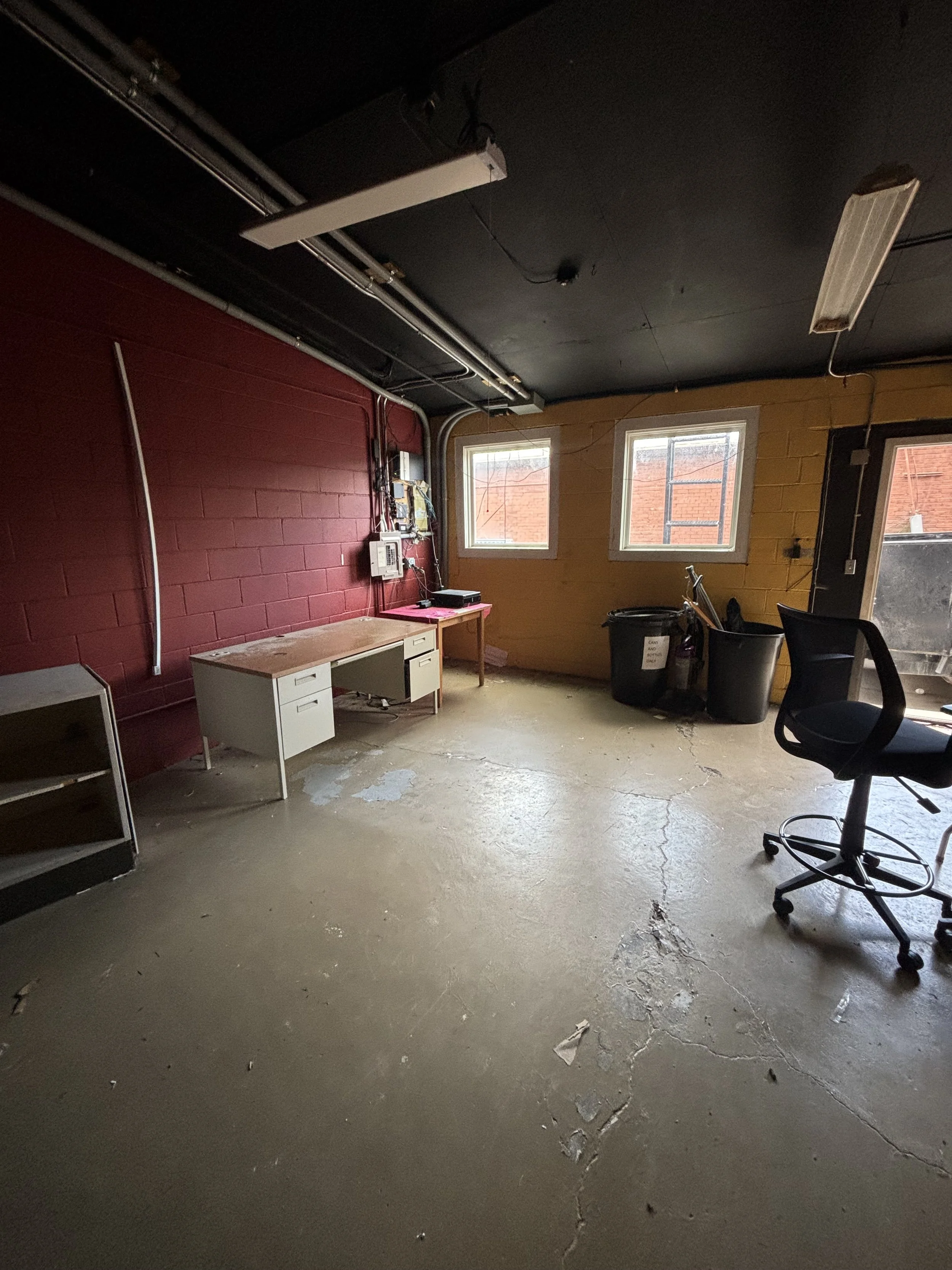 An unfinished indoor room with a red brick wall on the left and a yellow brick wall on the right. There are two windows on the yellow wall, a white desk with drawers, and a black office chair. The ceiling is black with visible lights, wiring, and a white rectangular object hanging.