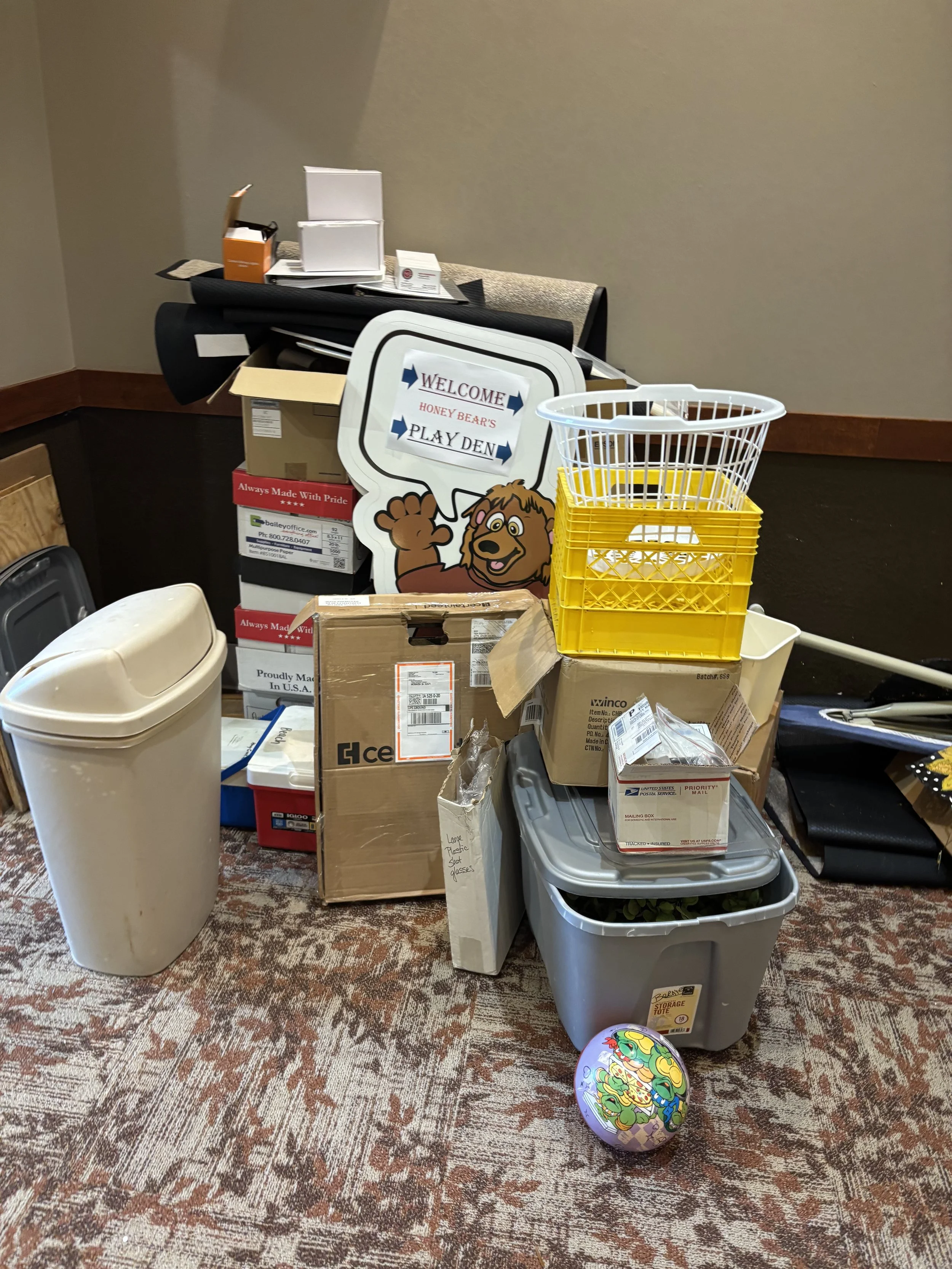 A cluttered room with assorted boxes, containers, and miscellaneous items on the carpeted floor and table. There is a decorative dragon-themed orb on the floor, a cardboard box, plastic storage bins, and a smiling cartoon lion sign with the text "WELCOME HONEY BEAR'S PLAY DEN."