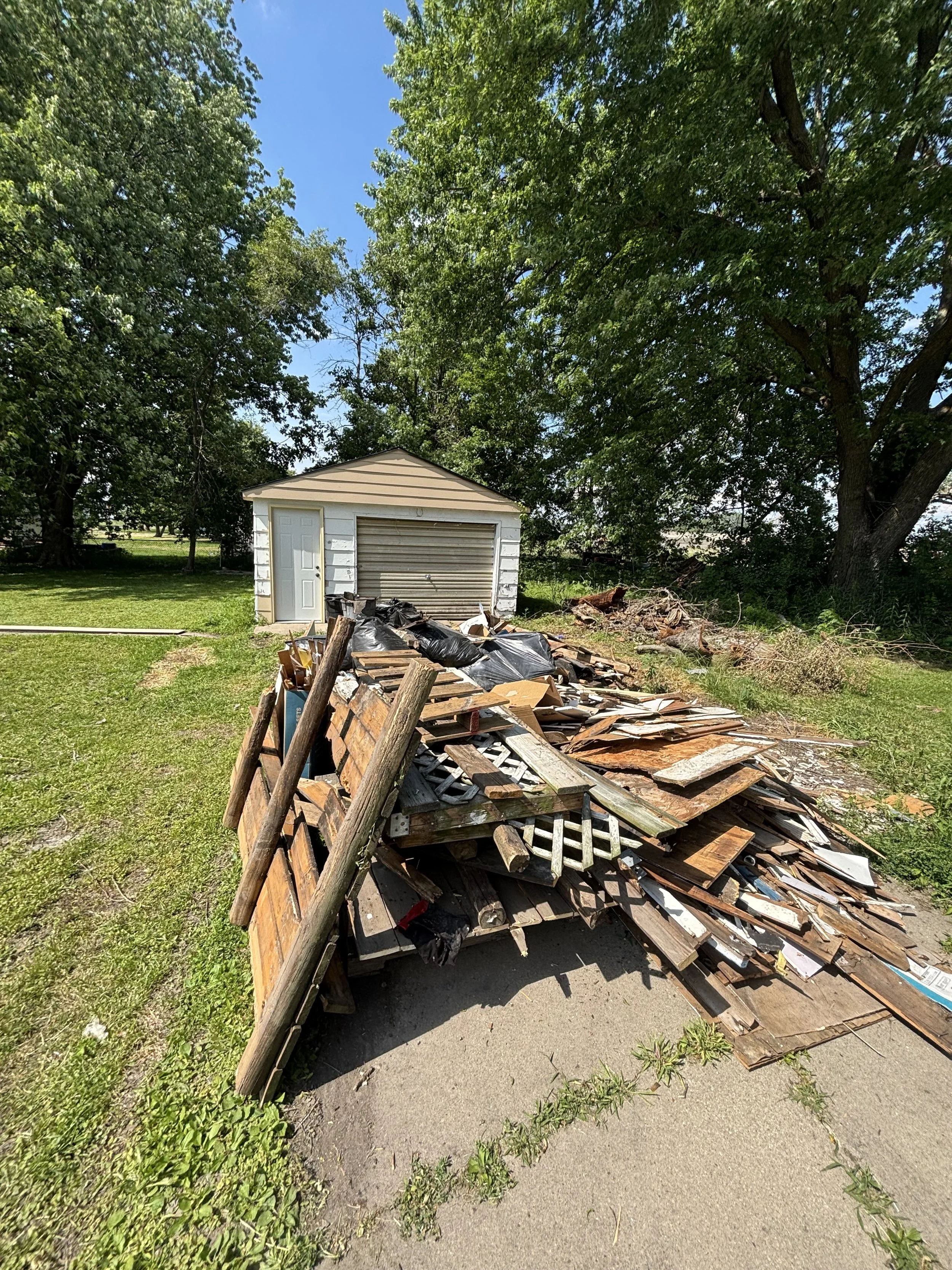 A small white shed with a beige roof and closed garage door standing in a grassy yard. In the foreground, there's a large pile of wooden debris, including old wooden panels, planks, and black trash bags. Tall trees with lush green leaves shade the area, and a bright blue sky is visible overhead.