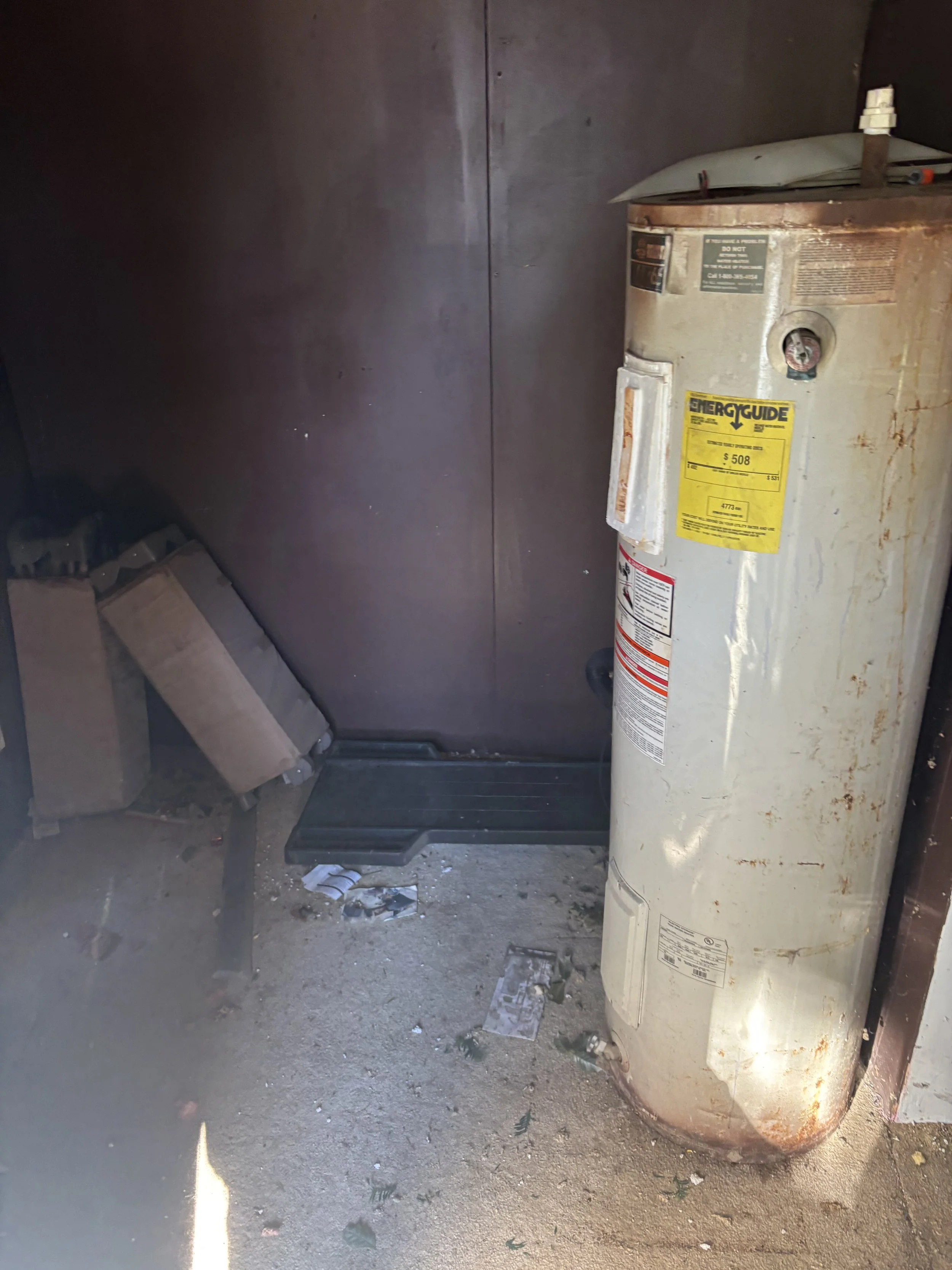 Old, rusty water heater with a yellow energy guide label, positioned next to a dark brown wall, on a dirty floor with scattered debris and some cardboard boxes.
