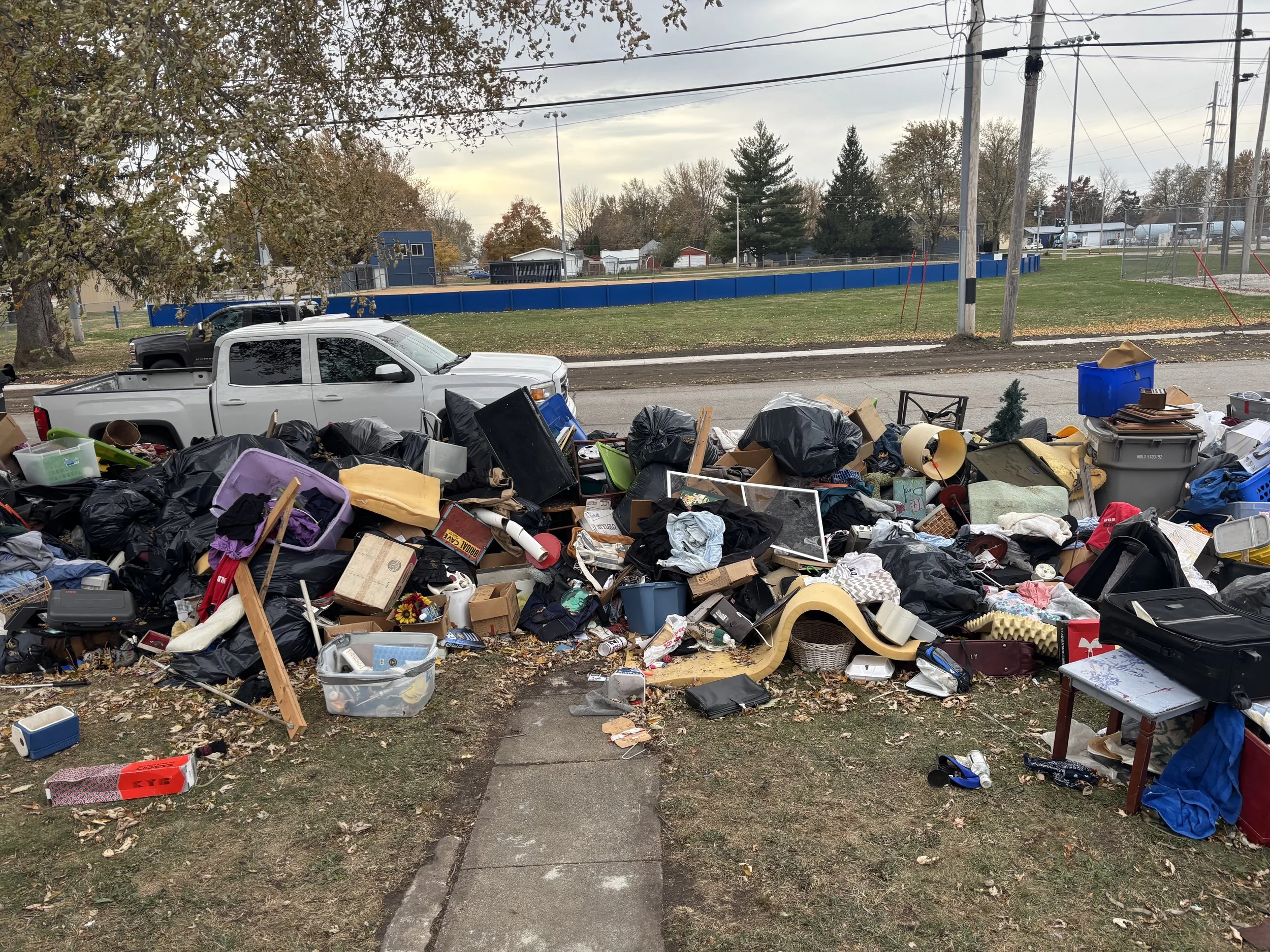 A large pile of assorted trash and discarded items spilling onto a sidewalk, with a white truck parked behind it, near a street with trees, utility poles, and a sports field or park in the background.