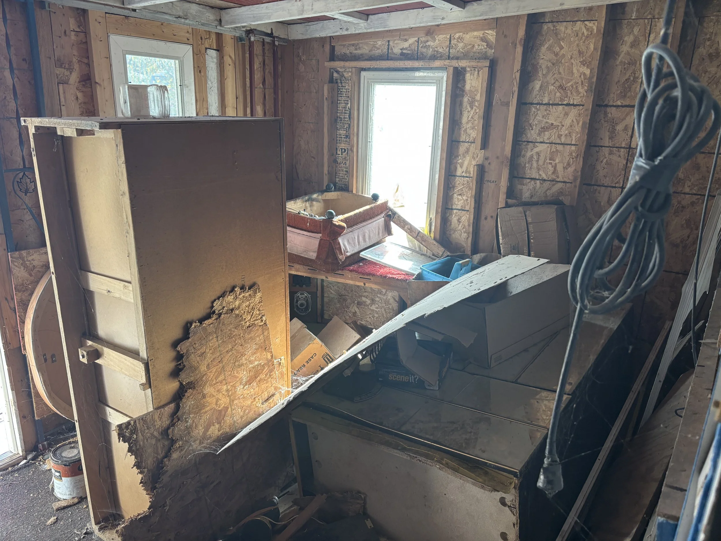 A cluttered workshop with exposed wooden walls, a small window, and a table partially covered with boxes and tools, indicating ongoing construction or renovation.