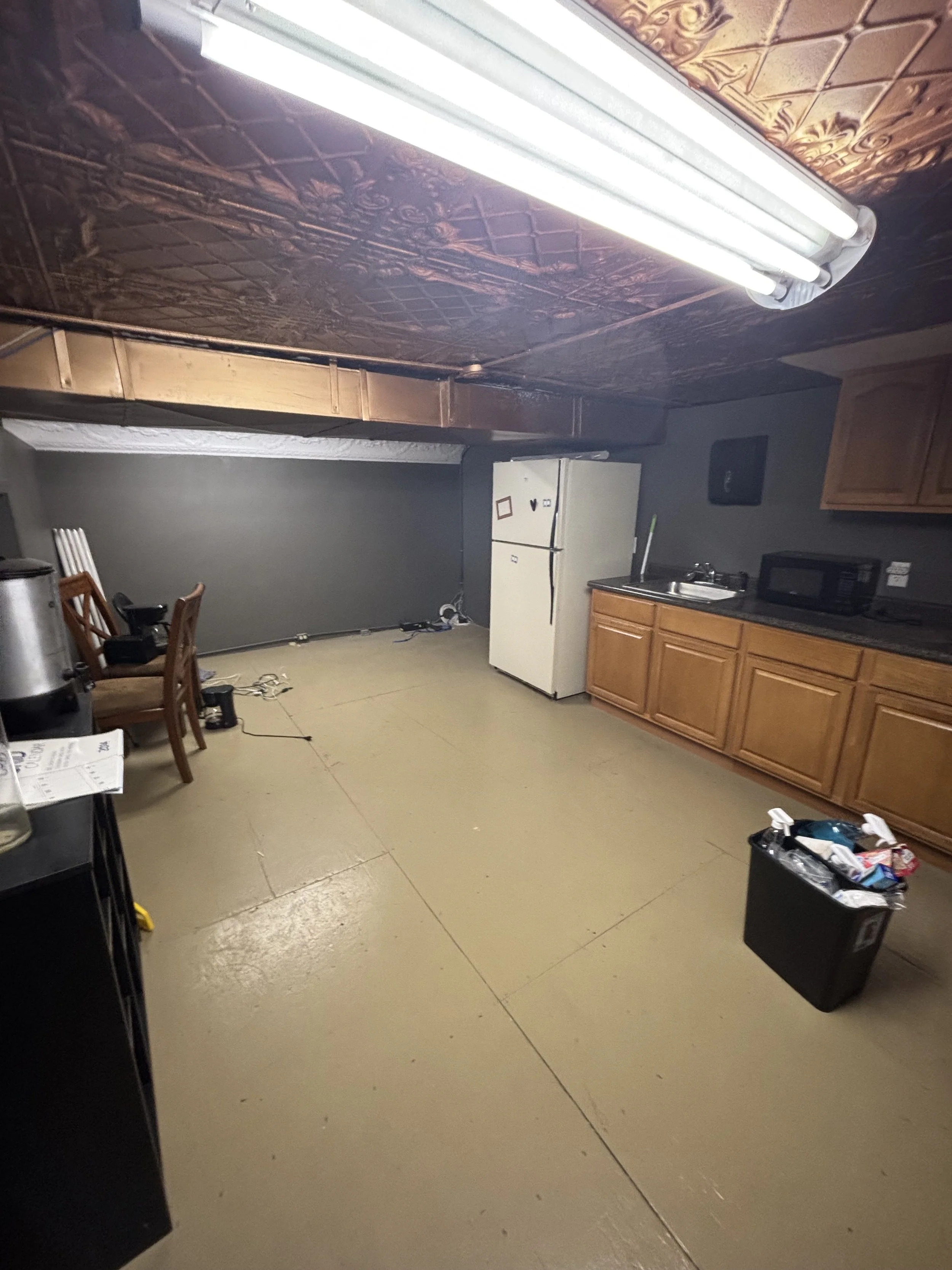 A kitchen with wooden cabinets, a white refrigerator, a microwave, a sink, a countertop, a black trash bin, and a fluorescent light on the ceiling. The floor is beige with visible tiles.
