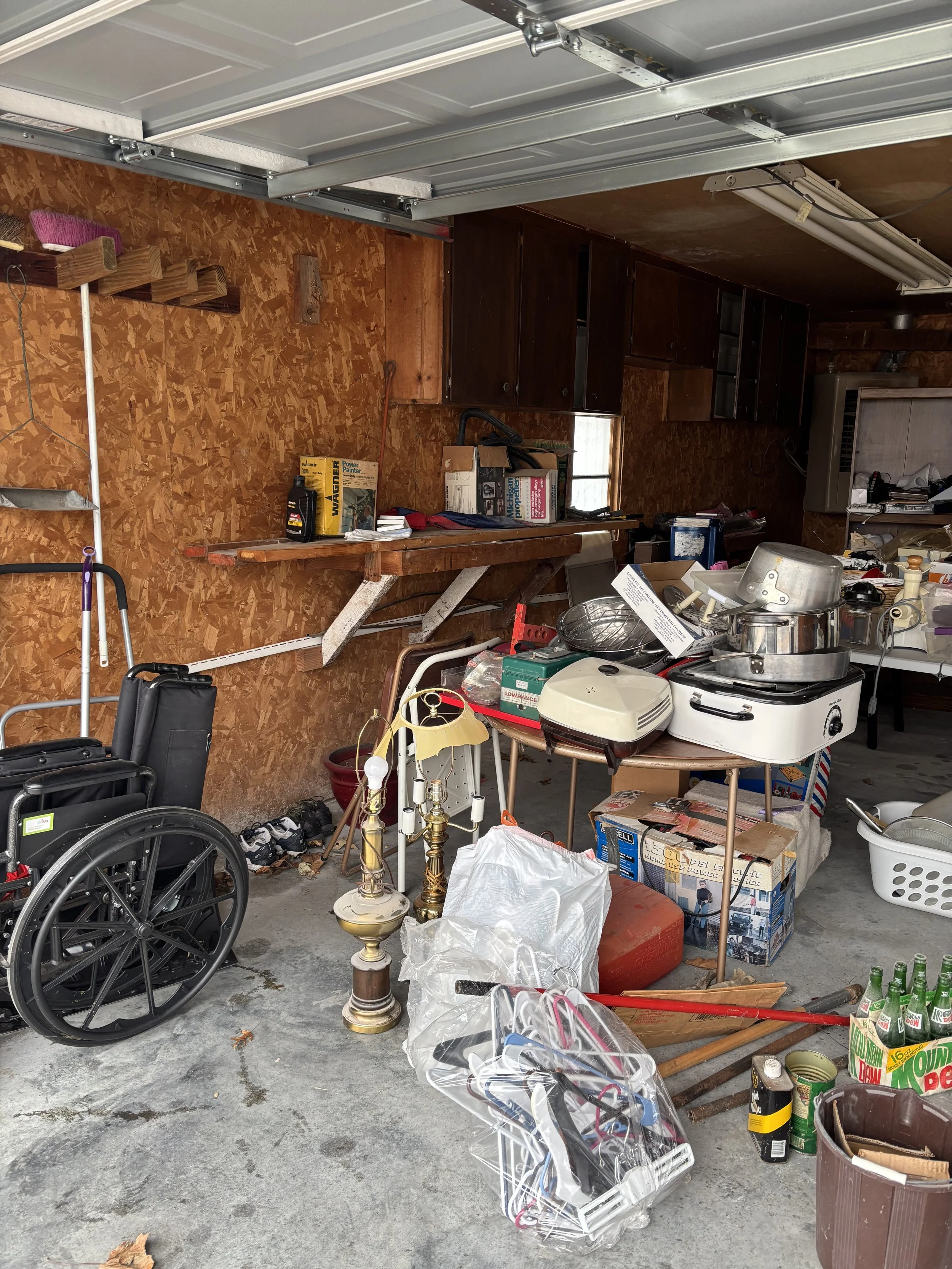 Garage cluttered with various household and tools items including a wheelchair, lamp, plastic bags, boxes, kitchen appliances, and miscellaneous clutter.