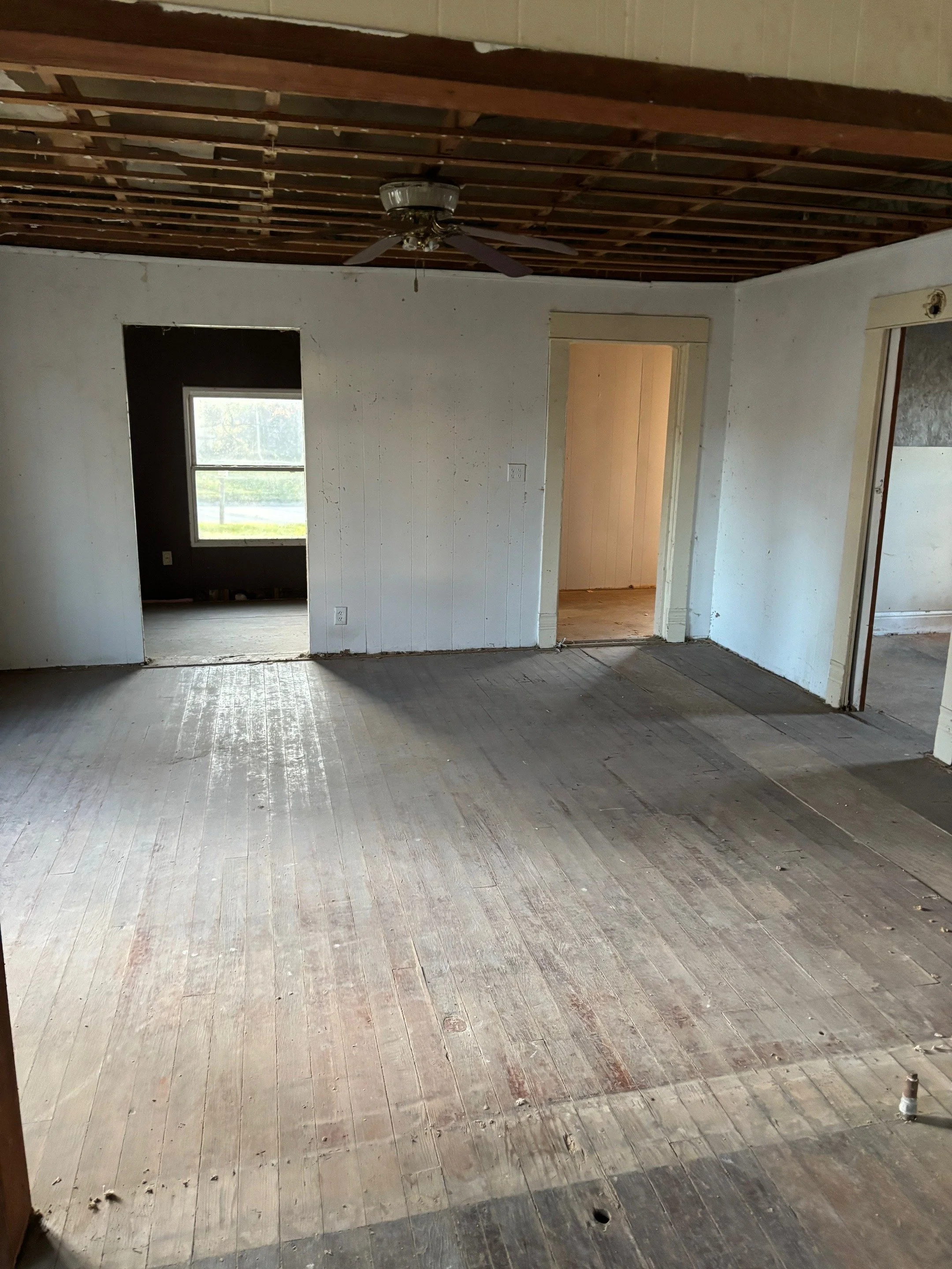 Empty living room with unfinished wooden floor, white walls, open doorways to other rooms, ceiling fan, and exposed ceiling beams.