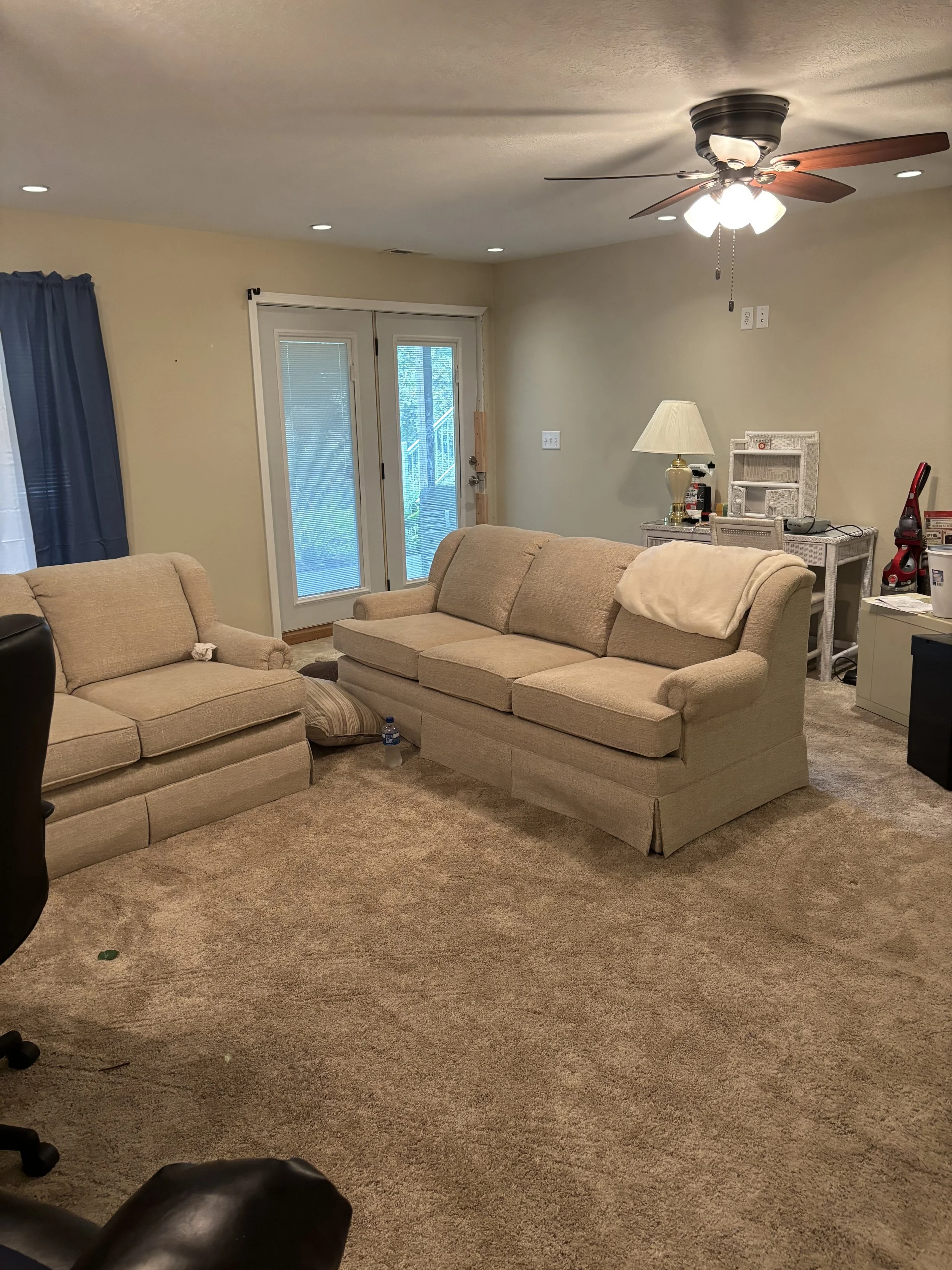 Living room with two beige sofas, a glass door with blue curtains, and a ceiling fan with lights. There is a white side table with a lamp, and a vacuum cleaner in the corner.