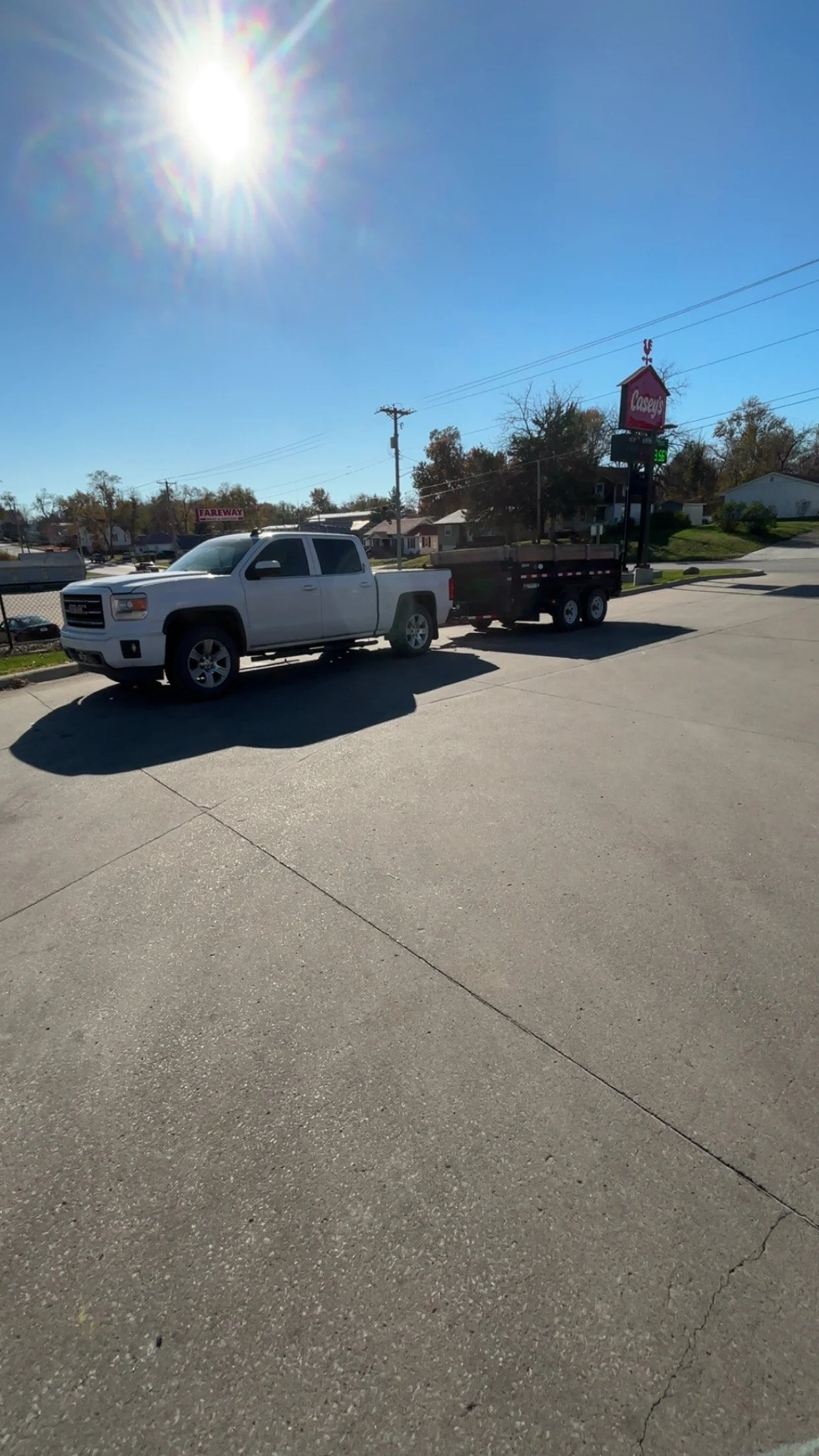 A white pickup truck with a trailer attached parked on a parking lot on a sunny day. In the background, a sign for Casey's General Store and a few houses are visible.