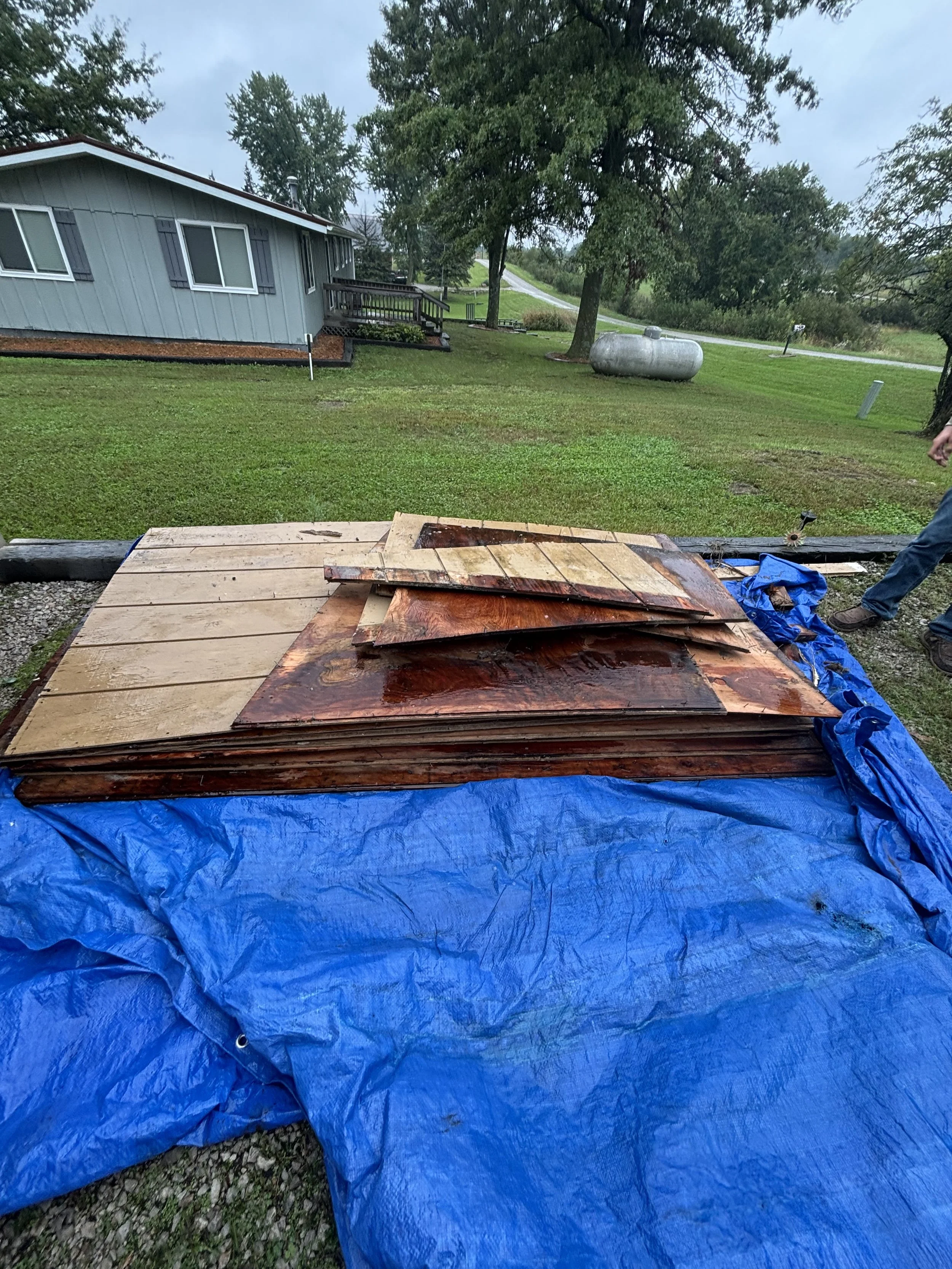 A pile of wooden planks and stained wood pieces on a blue tarp outdoors in front of a house with trees, grass, and a propane tank.