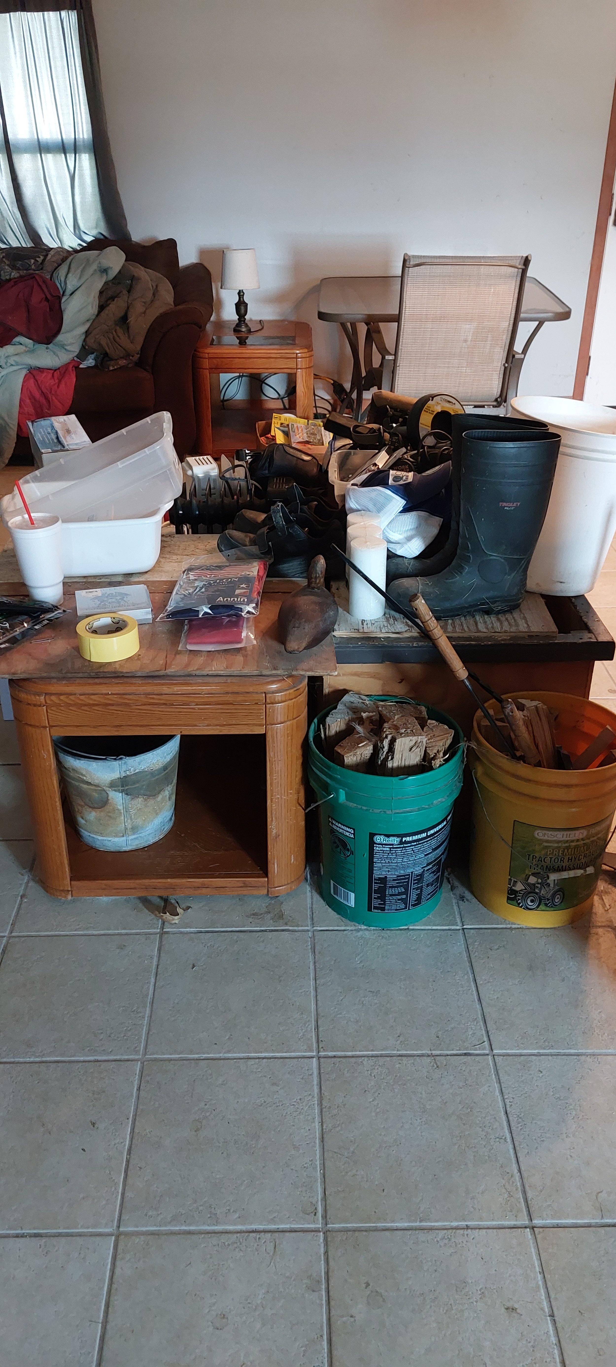 Living room cluttered with various objects including a table with tools, buckets, containers, a rubber boot, and other miscellaneous items.