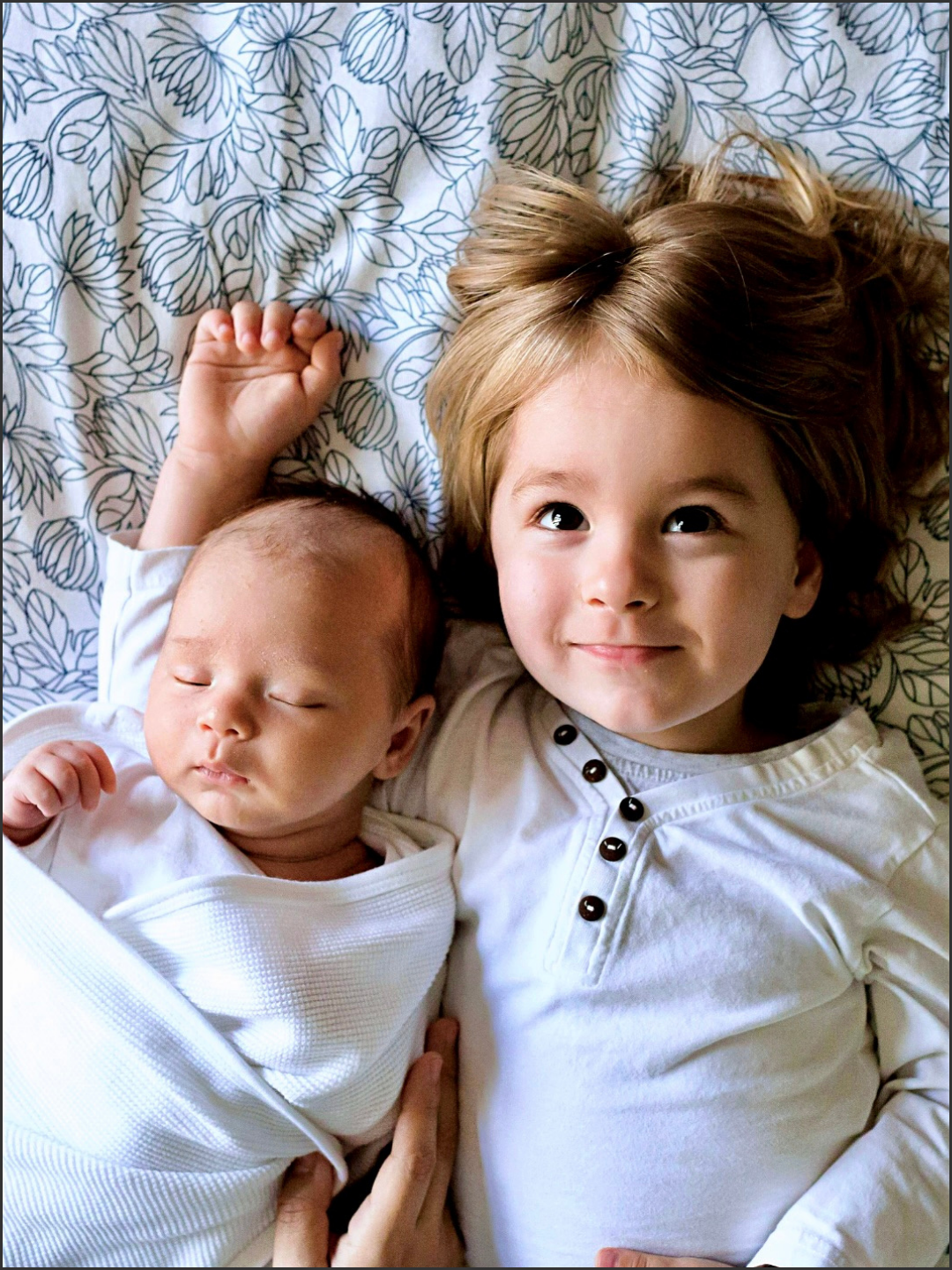 A young girl with long, wavy brown hair lying next to a sleeping baby with a patterned bedsheet in the background.