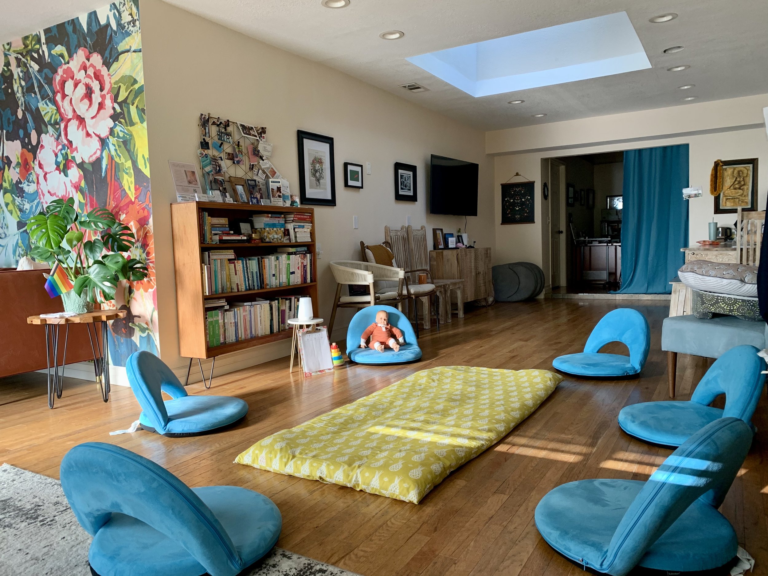 A cozy living room with blue papasan chairs arranged around a yellow mat on the wooden floor. There is a large bookshelf filled with books, artwork on the wall, a crib with a doll, and a skylight in the ceiling providing natural light.