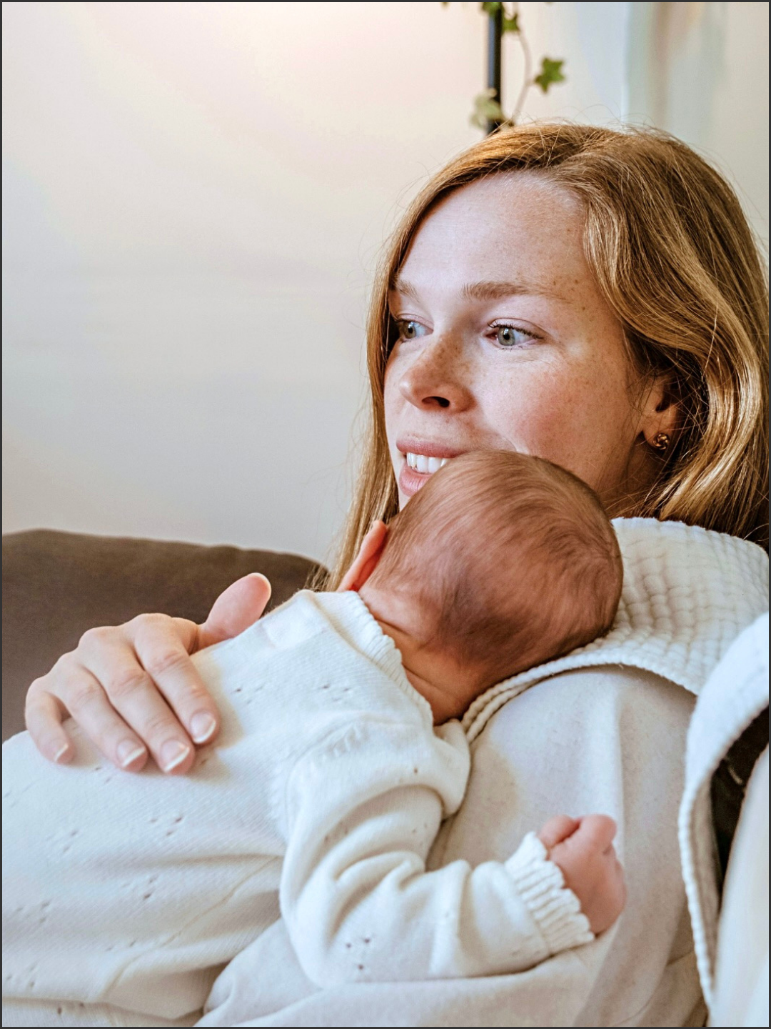 A woman with red hair holding a baby close to her chest, with the baby resting against her shoulder. The woman is looking out with a gentle expression, and the baby has their head turned to the side, showing soft hair and a white outfit.