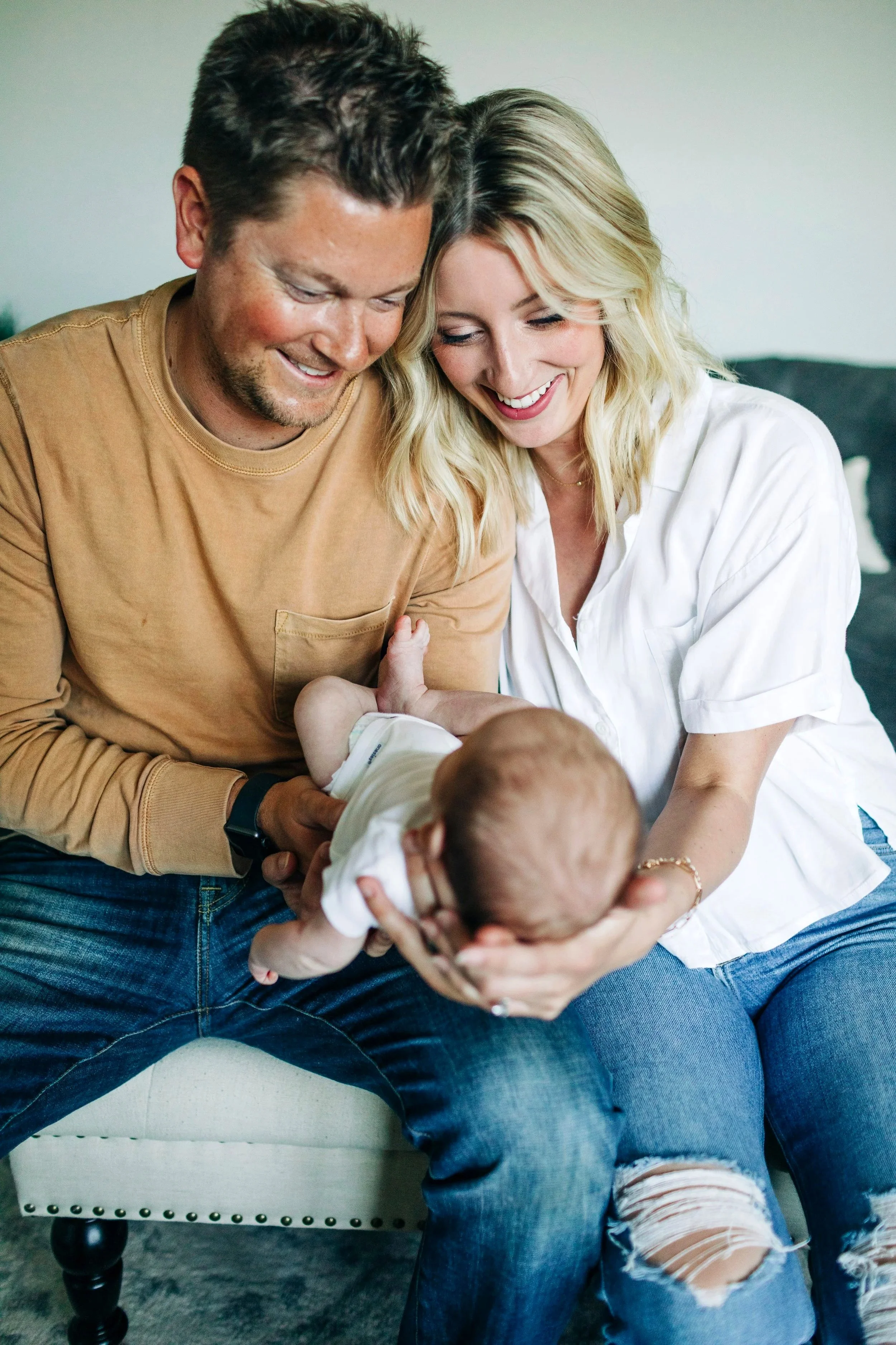 A smiling couple sitting on a white chair with a baby, holding and playing with the baby, in a well-lit room.