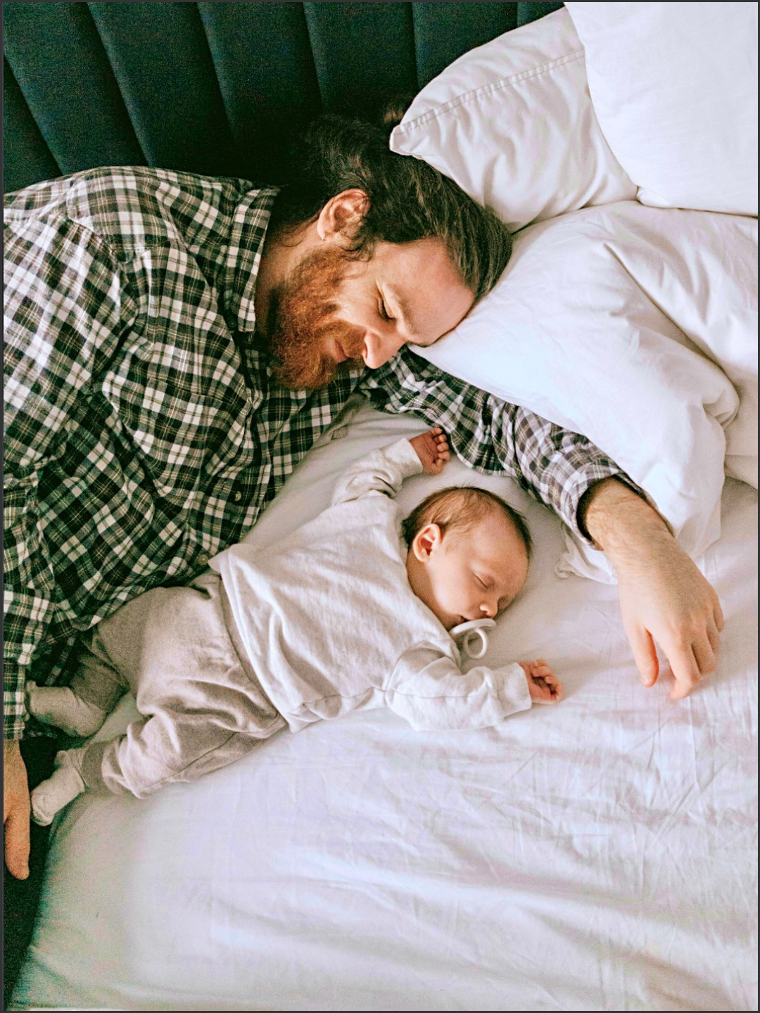 A man with a beard and long hair sleeping on a bed next to a young child, both lying on white sheets, with the man's arm extended across the bed and the child's hand resting near his face.