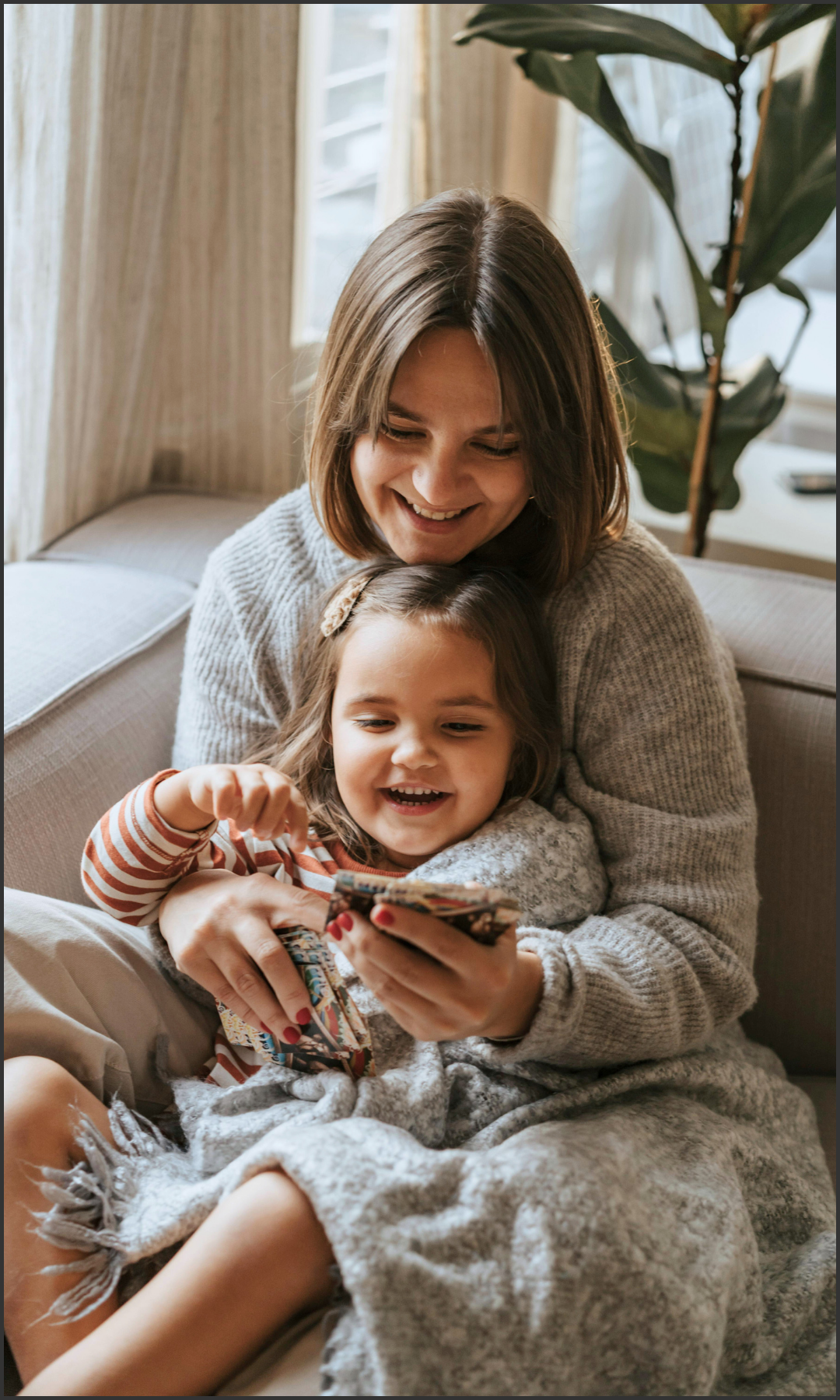A woman and a young girl sitting on a couch, looking at and smiling at a phone together in a cozy, well-lit room with a large potted plant and curtains in the background.