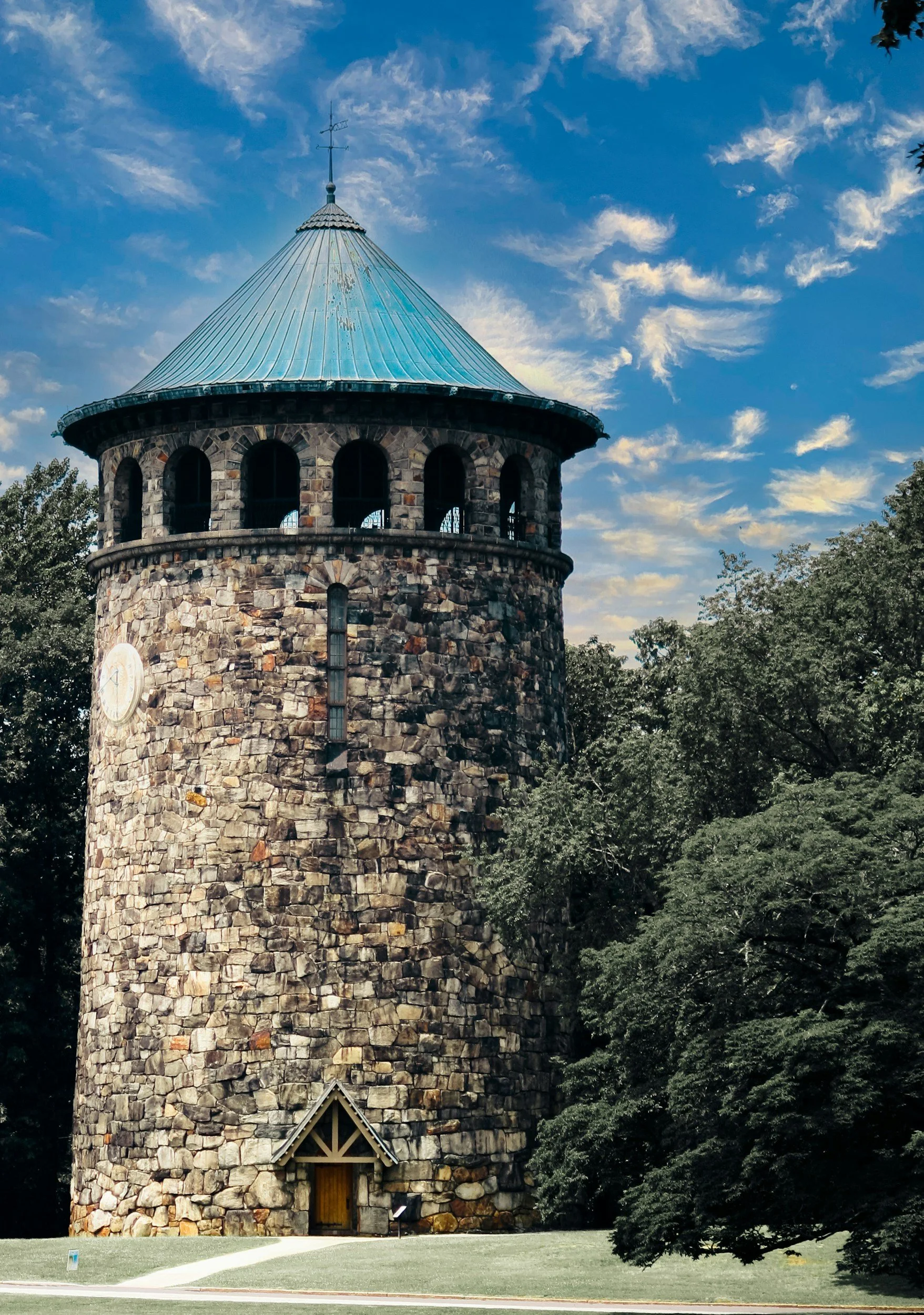 Stone tower with a conical metal roof, arched windows near the top, and a small wooden door at the base, surrounded by trees and a grassy area under a blue sky with scattered clouds. Project Elevate 360. Delaware.