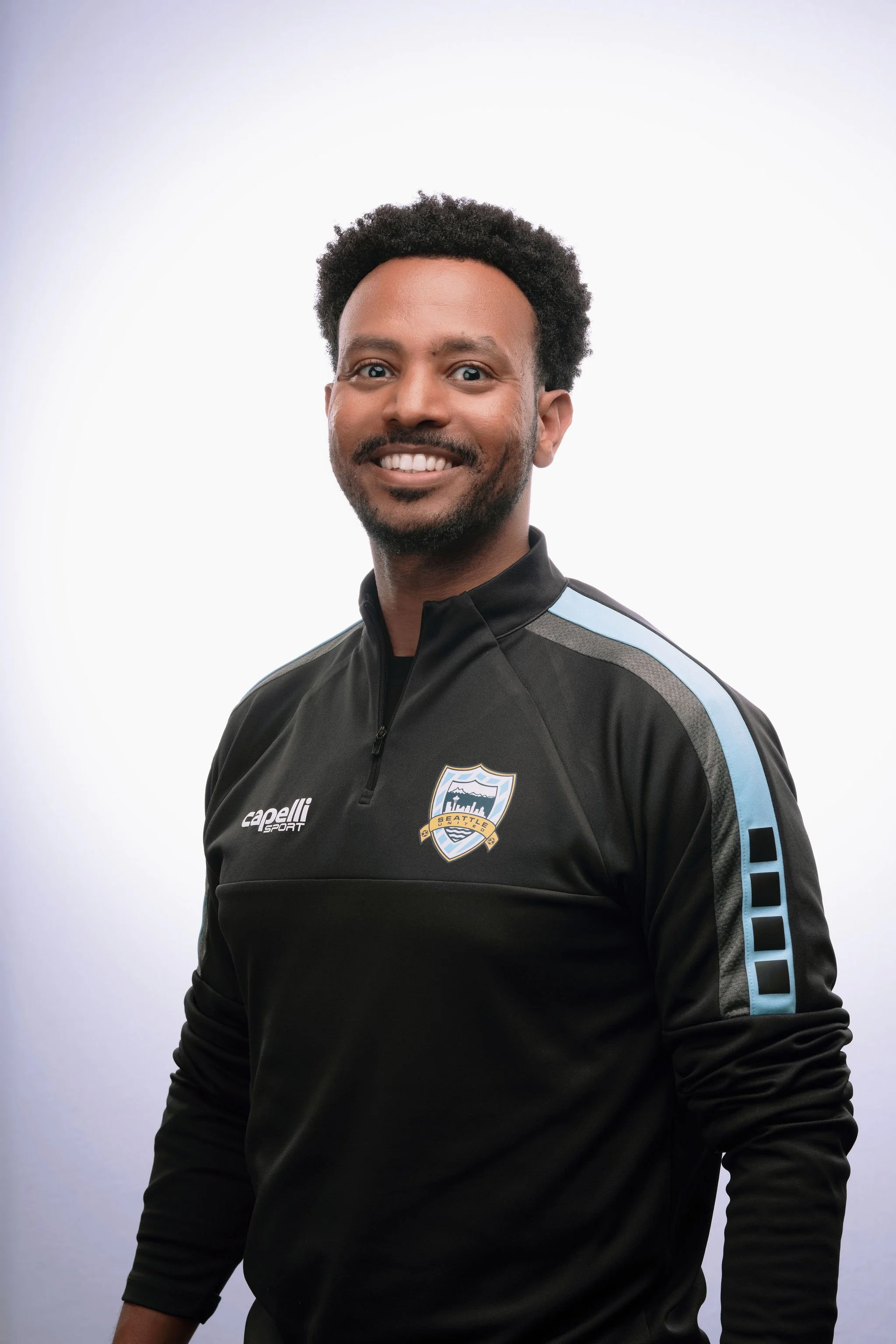 A smiling man with short curly hair and a beard, wearing a black sports jacket with a Seattle soccer team logo, standing against a plain white background.