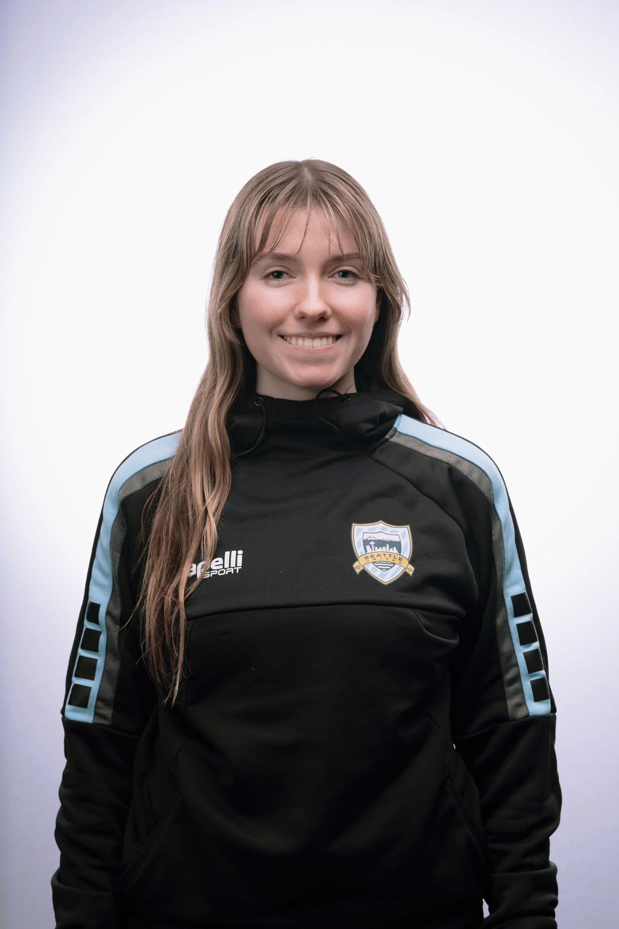 A young woman smiling, wearing a black sports jacket with Seattle Sounders Soccer Club logo on the chest, against a plain white background.